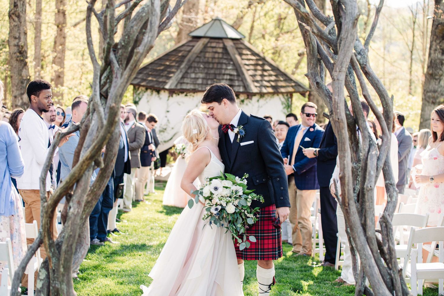 Bride and groom share a kiss at outdoor wedding ceremony, groom in tartan kilt, bride holding white bouquet.