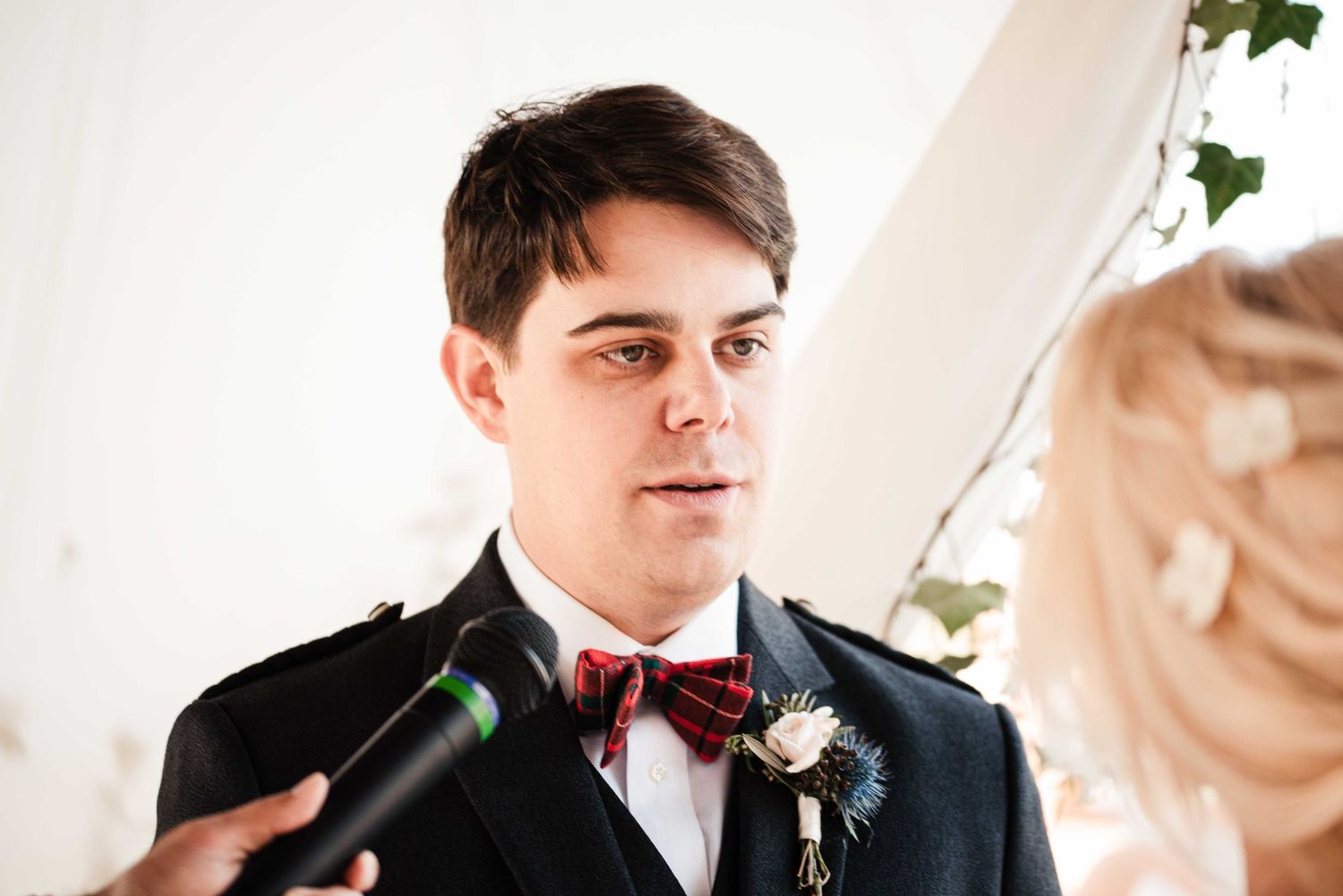 Groom in black suit with red tartan bow tie and thistle boutonniere speaks vows at wedding ceremony.
