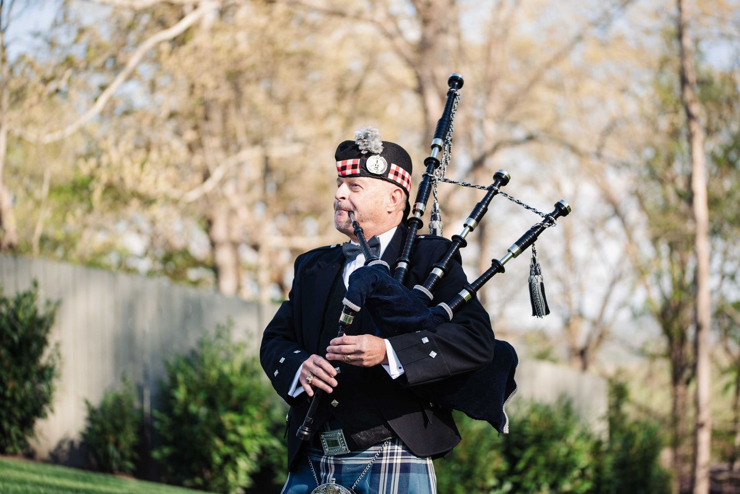 A bagpiper in traditional Scottish attire plays bagpipes outdoors among trees.