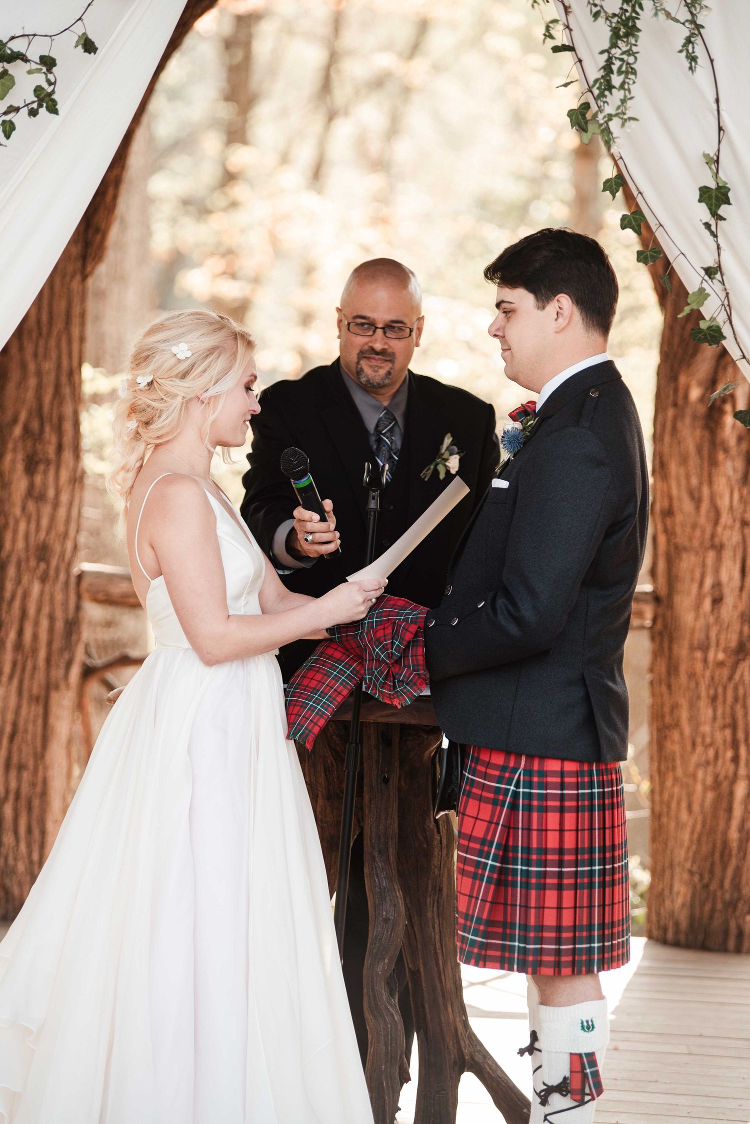 Bride reads vows to groom wearing red tartan kilt at outdoor wedding ceremony with officiant.