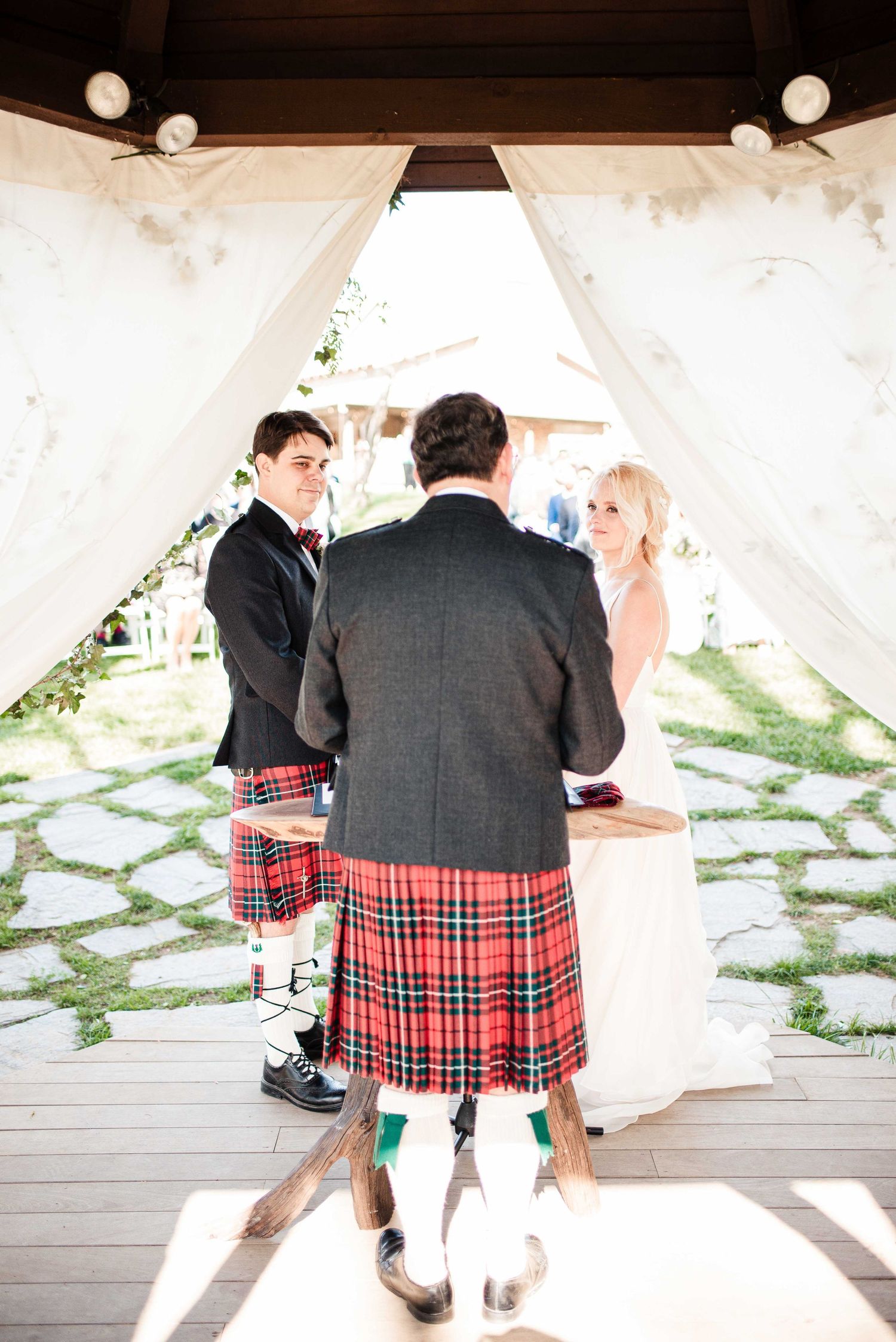 Outdoor wedding ceremony with groom in kilt, bride in white dress, and officiant under draped gazebo.