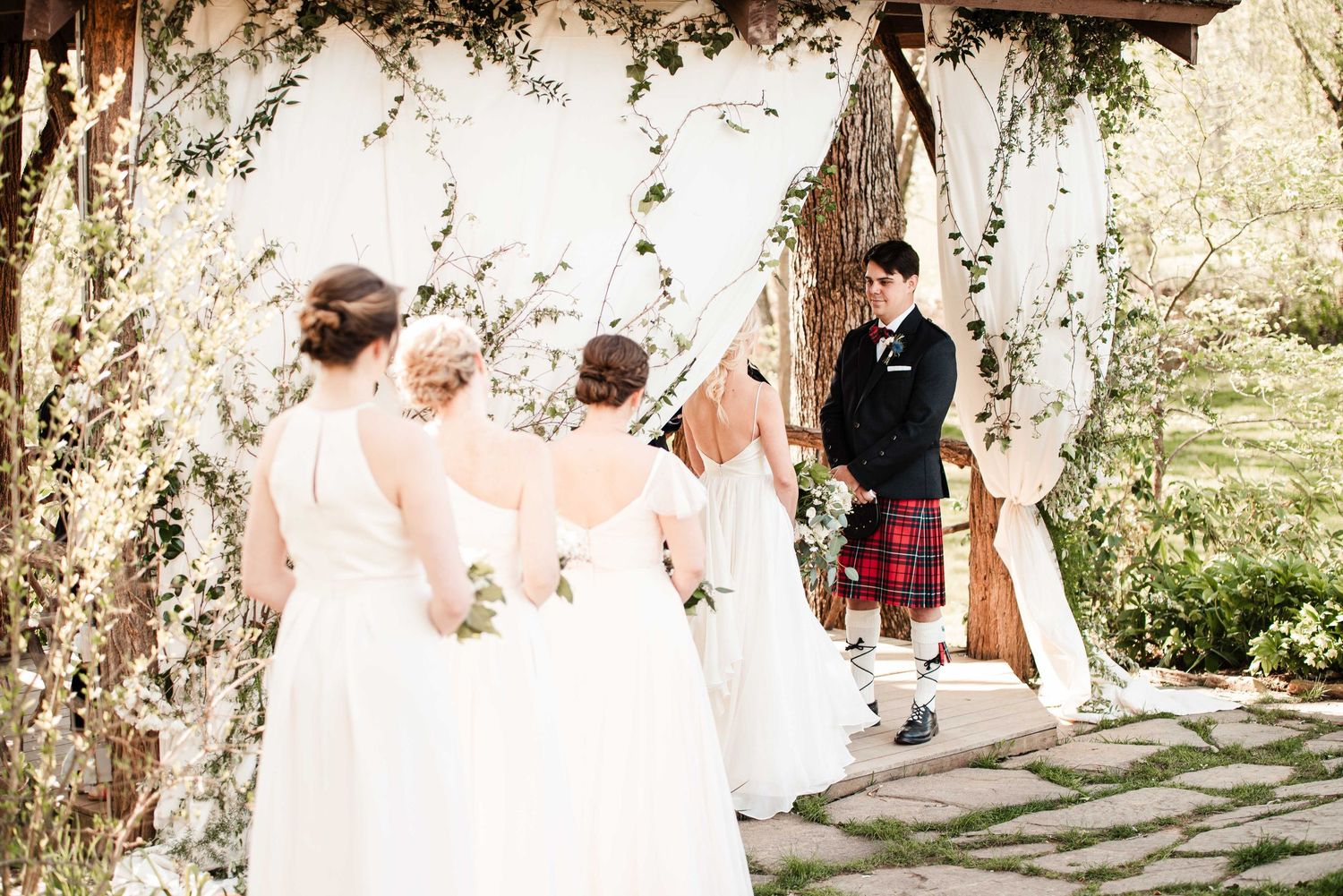 Bridesmaids in white gowns walk toward a groom in a Scottish kilt at an outdoor floral wedding ceremony altar.