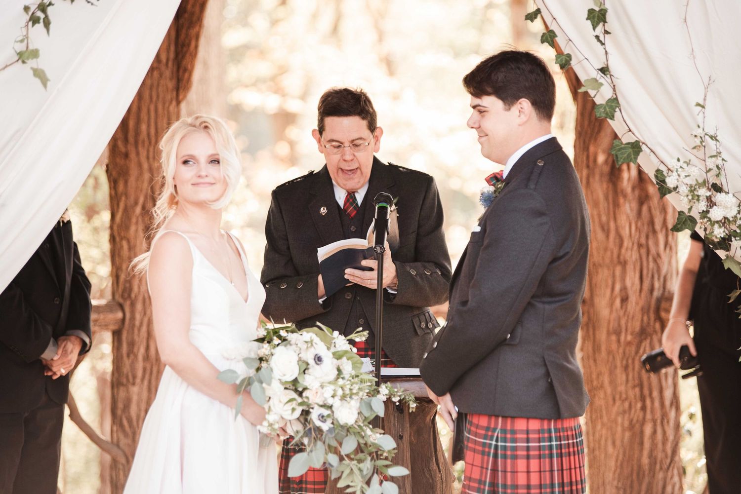 Bride and groom exchange vows outdoors under a floral arch with an officiant at a woodland wedding ceremony.