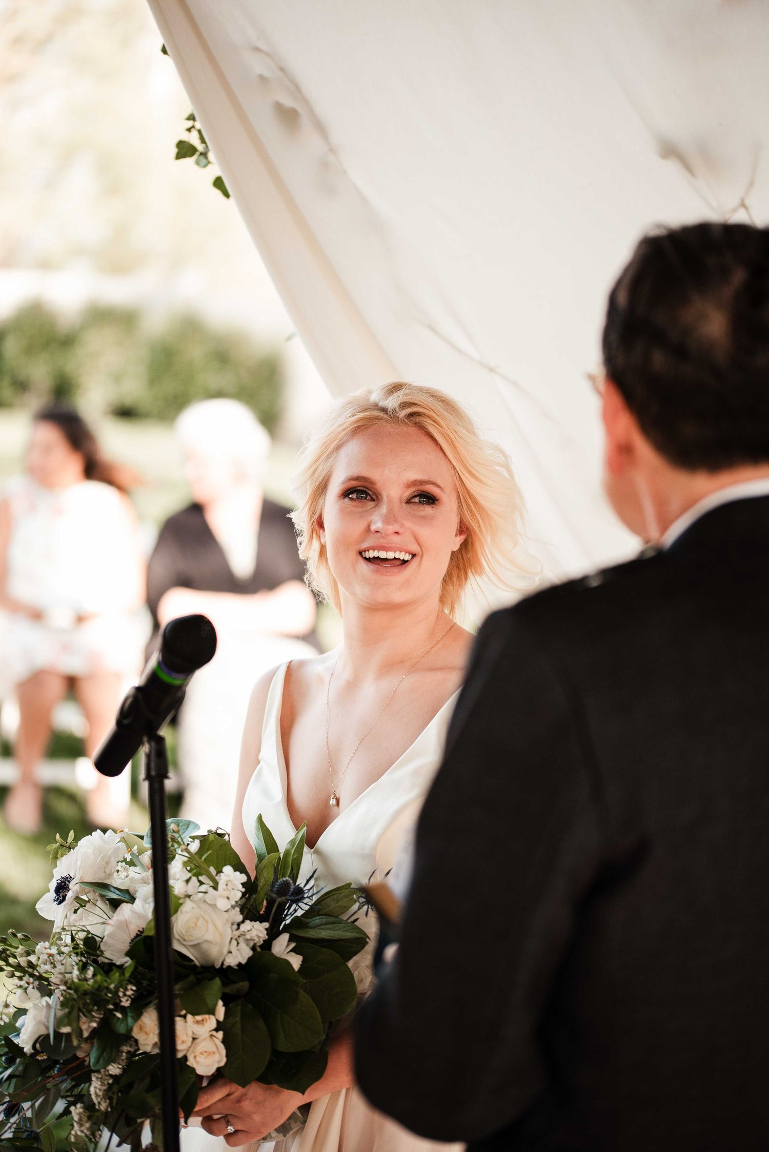 Smiling blonde bride holding white floral bouquet during outdoor wedding ceremony under white tent.