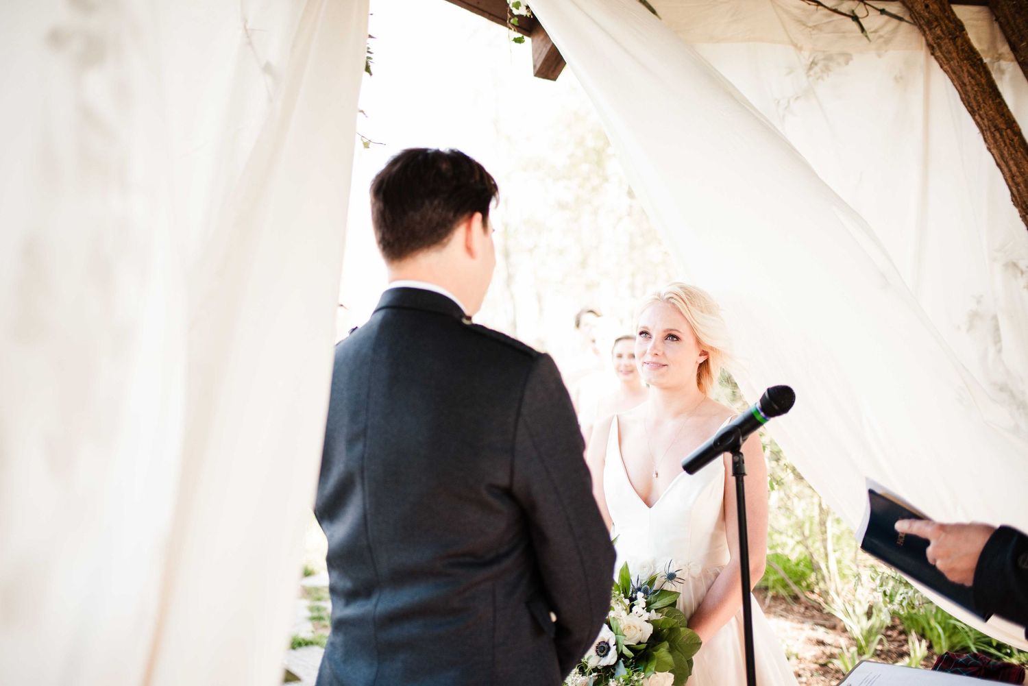 Bride and groom exchange vows at outdoor wedding ceremony under white draped canopy with floral bouquet.