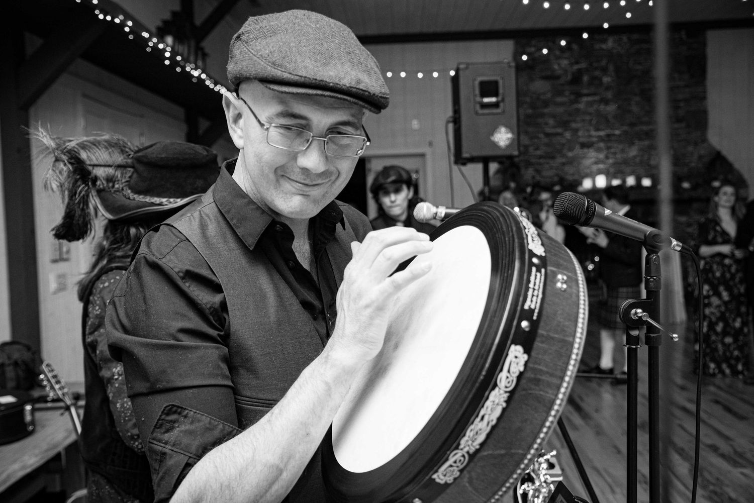 Black and white photo of a man wearing a flat cap and glasses playing a bodhran drum at a live music event.