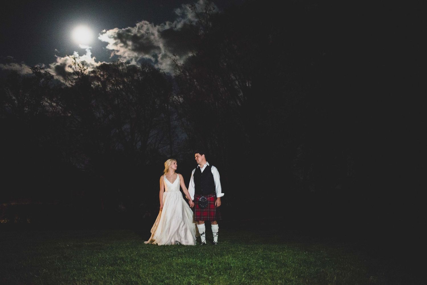 Bride in white gown and groom in red tartan kilt hold hands under moonlit night sky at outdoor wedding.