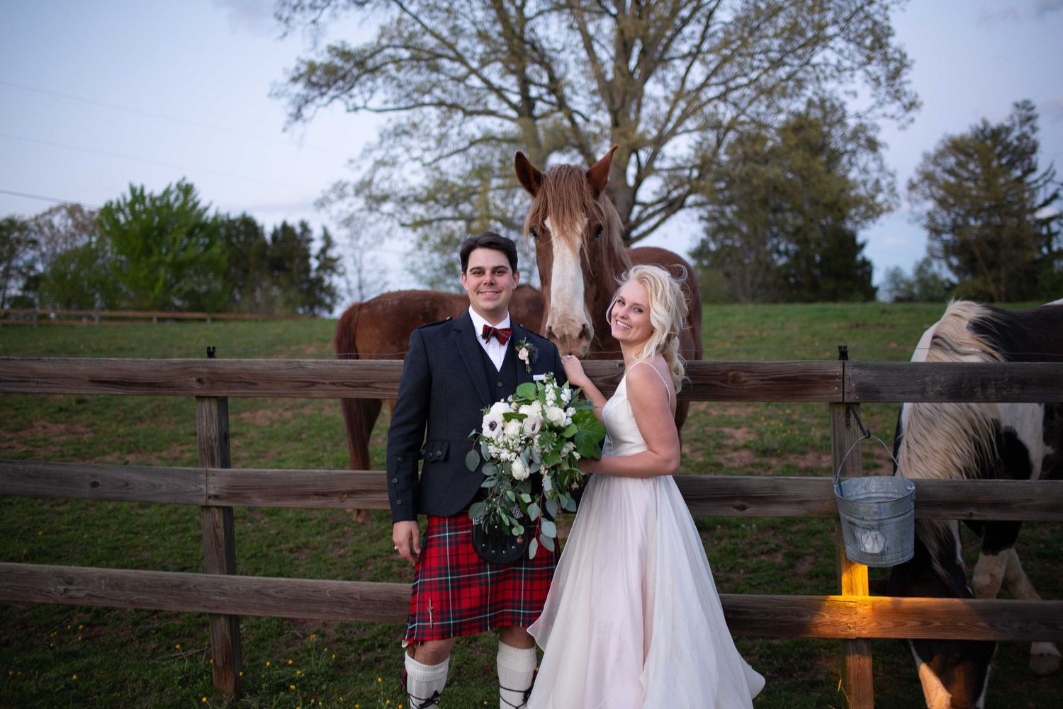 Bride and groom pose with horses at a rustic farm wedding, groom wearing a red tartan kilt, bride holding white bouquet.