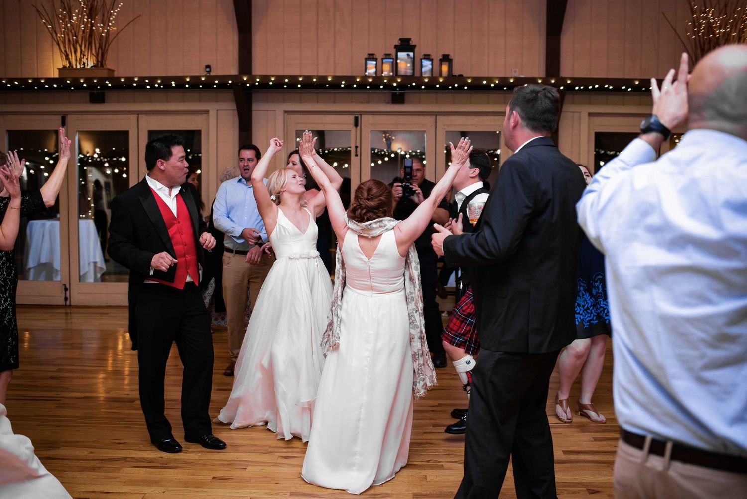 Two brides in white gowns dance joyfully with guests at an elegant wedding reception venue.