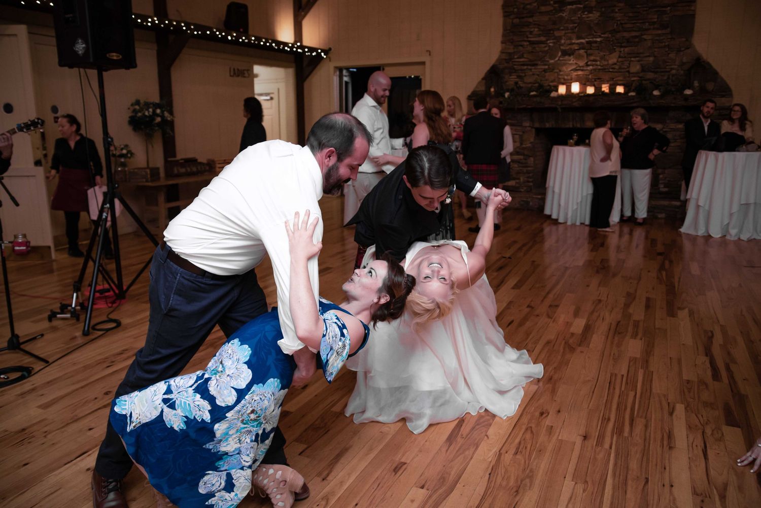 Guests dancing and dipping at a wedding reception on a wooden dance floor near a stone fireplace.