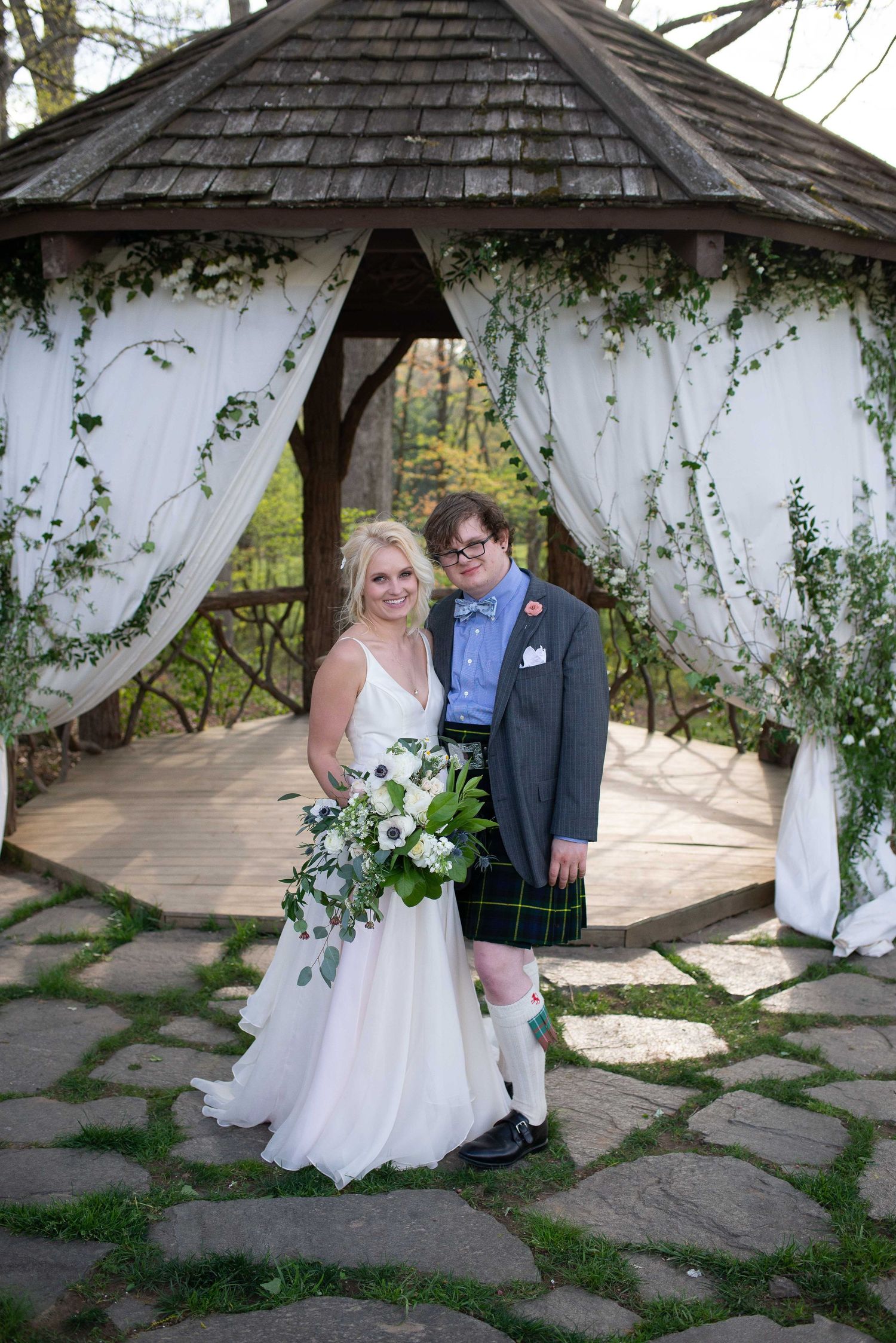 Couple posing under a floral gazebo, bride in white gown with bouquet, groom in gray suit and tartan kilt.