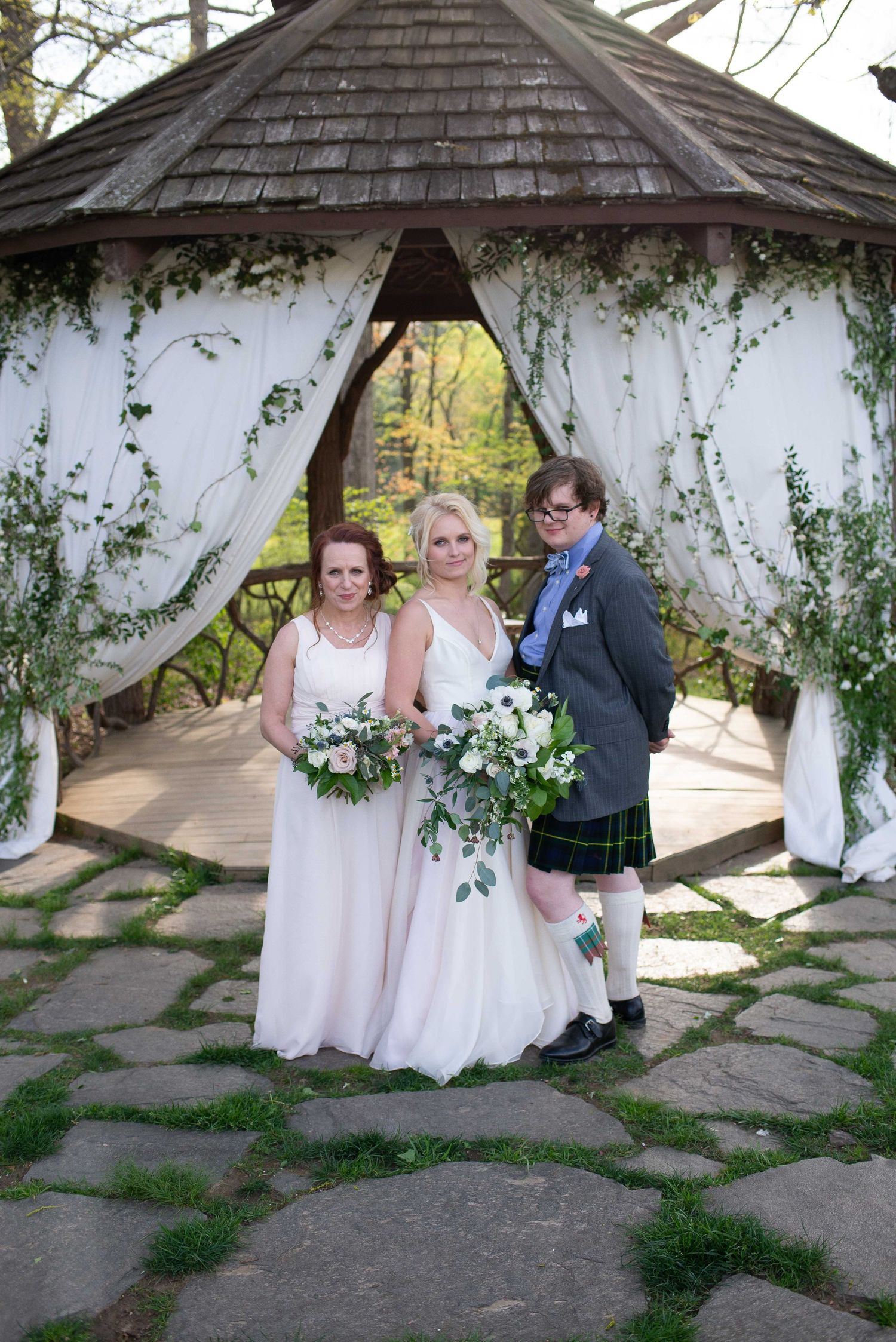 Two brides and partner pose under a floral gazebo with white drapes at an outdoor wedding ceremony.