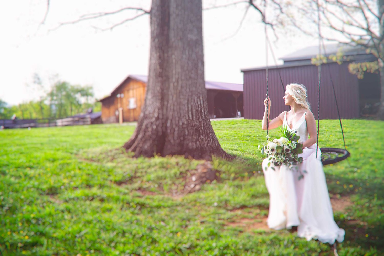 Bride in white gown holding bouquet stands by tire swing under large tree at rustic barn venue.