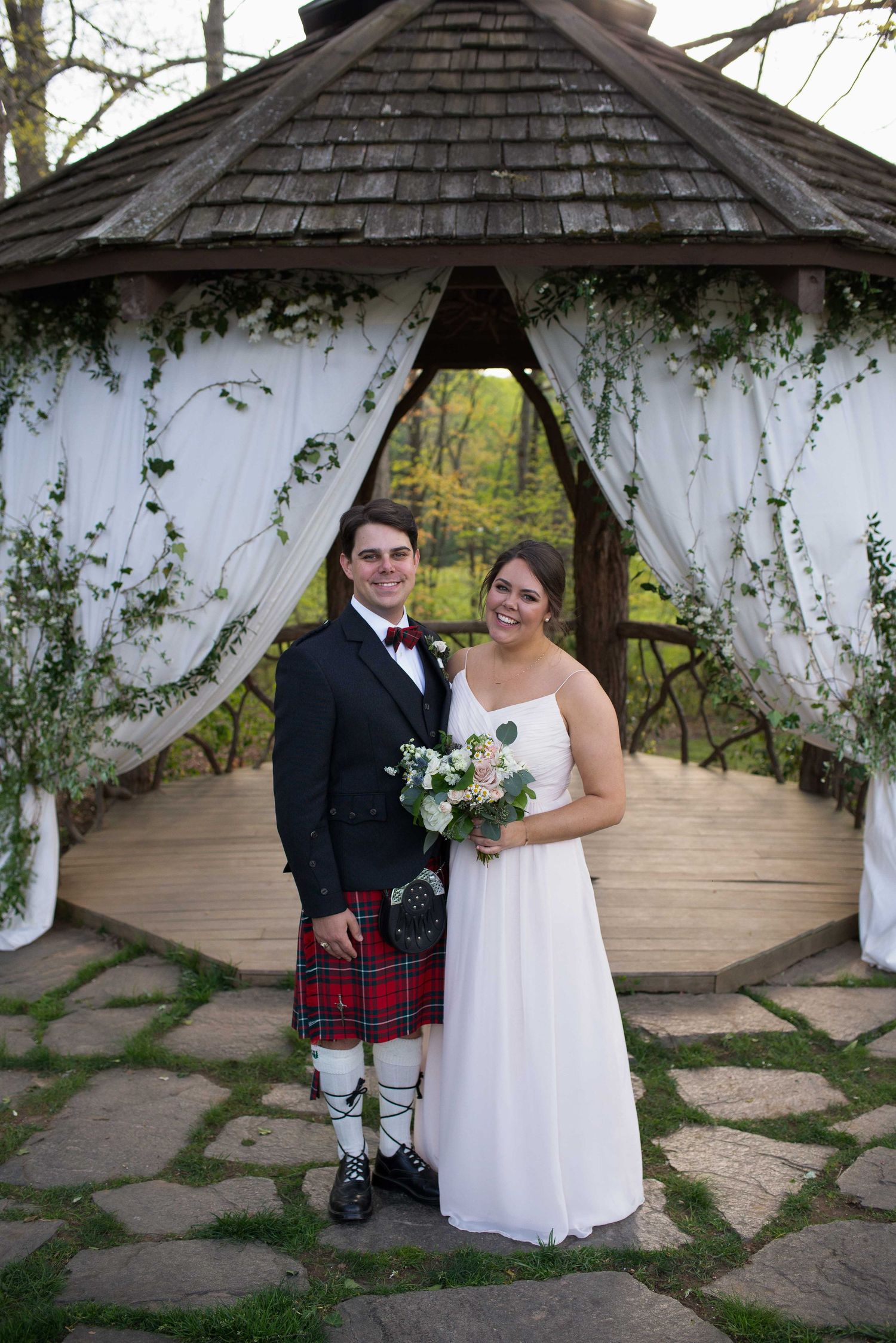 Couple poses under decorated gazebo, groom in red tartan kilt and bride in white gown holding bouquet, outdoor wedding ceremony.
