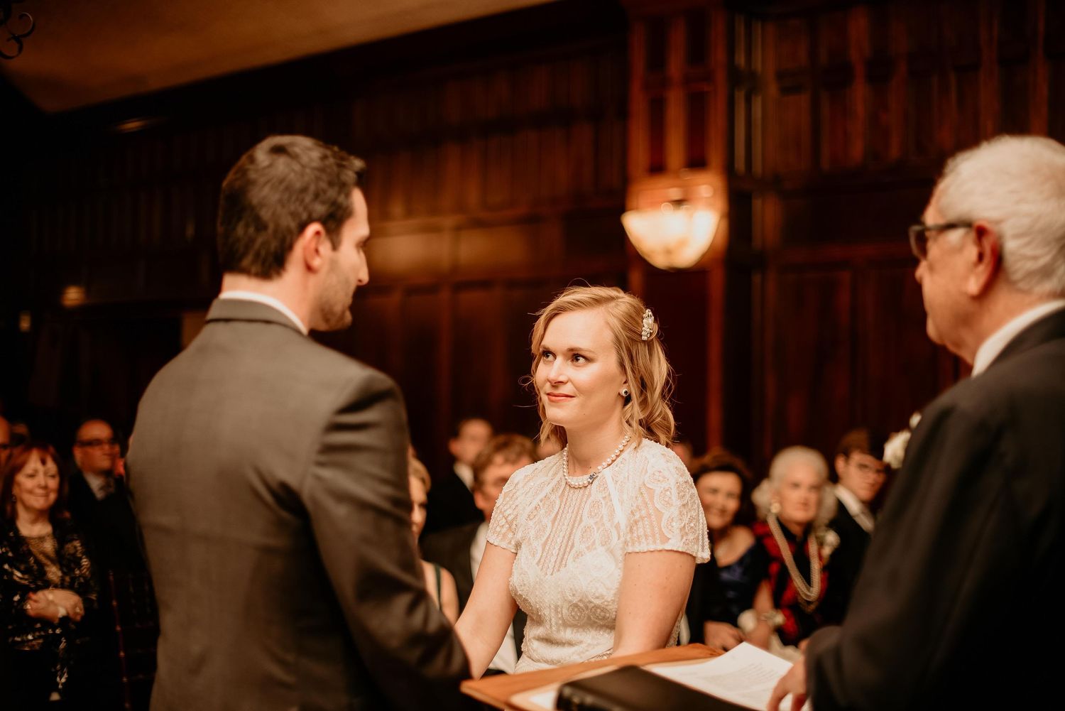 Bride smiling at groom during wedding ceremony in elegant wood-paneled venue with guests in background.