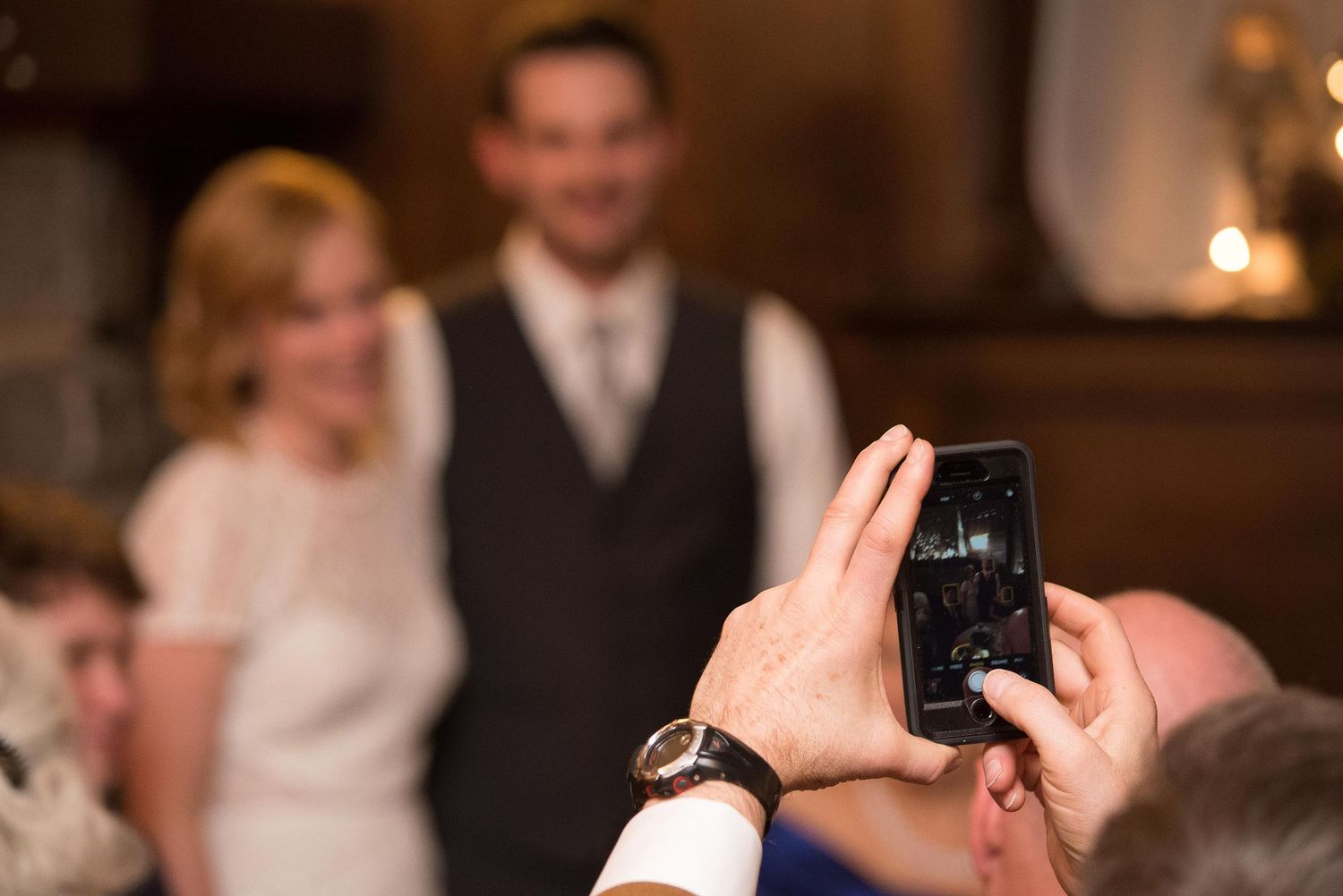 Person photographing a smiling bride and groom with a smartphone at a wedding reception indoors.