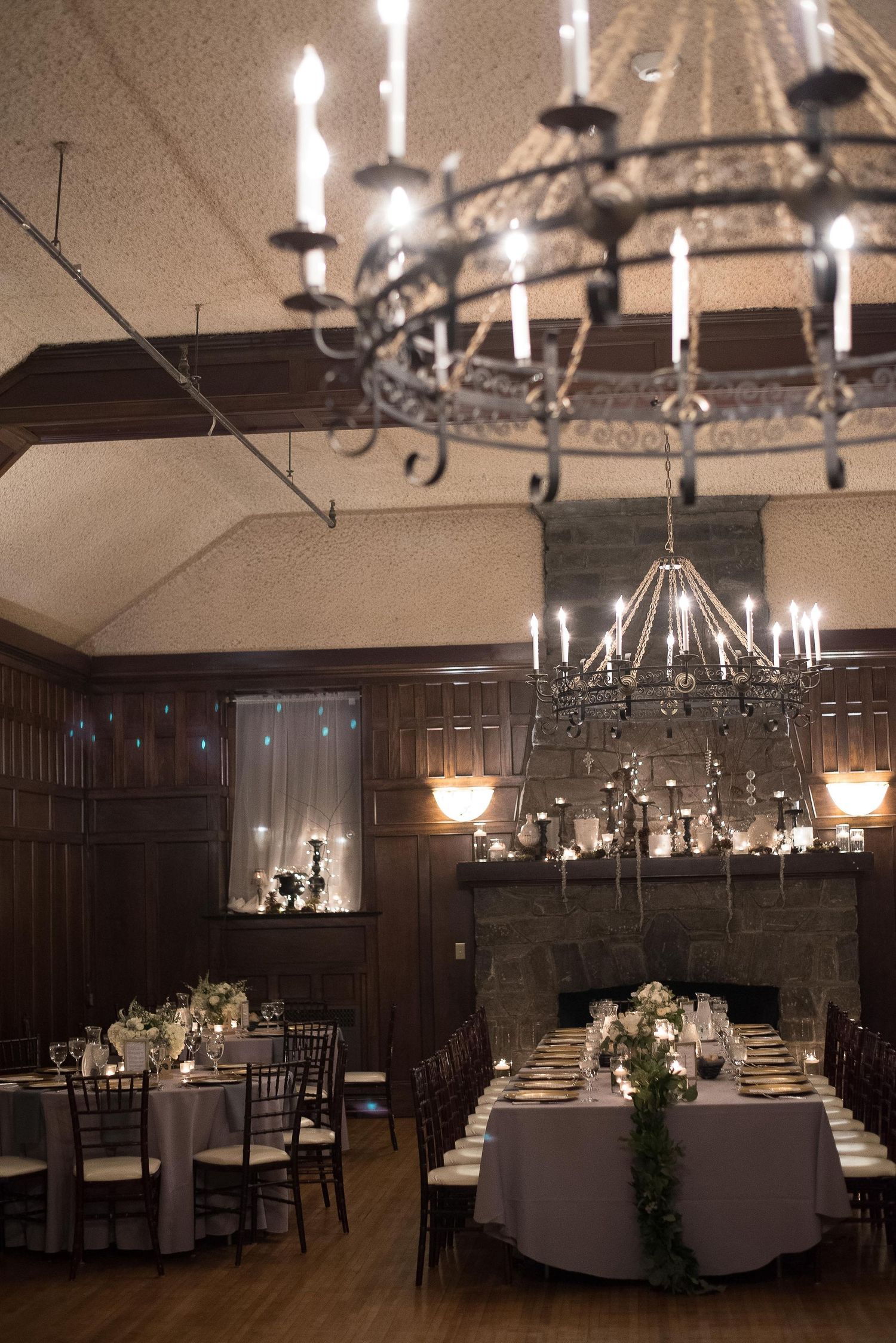 Elegant wedding reception hall with ornate iron chandeliers, white-draped tables, greenery centerpieces, and a stone fireplace.