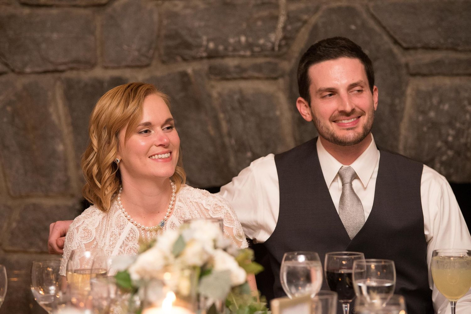 Smiling couple at a wedding reception table with floral centerpiece and stone wall backdrop.