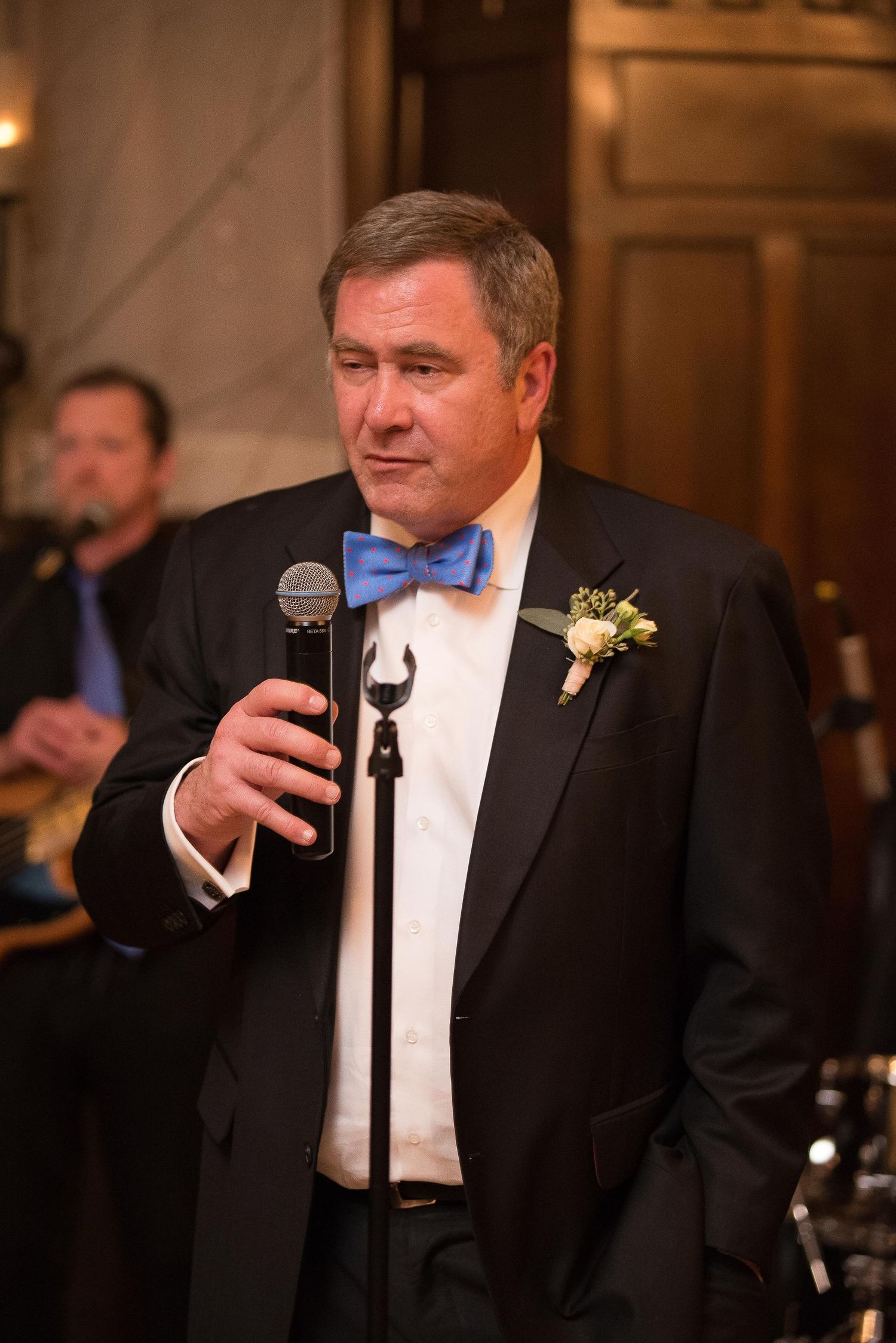 Man in tuxedo with blue bow tie speaks at microphone during wedding reception.