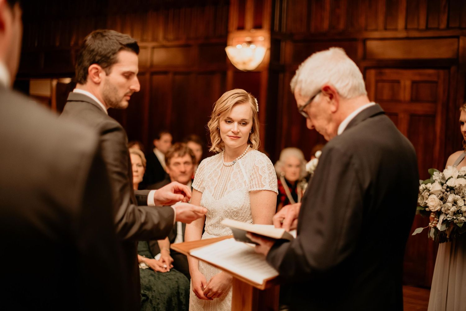Bride and groom exchanging rings during indoor wedding ceremony with officiant reading from book.