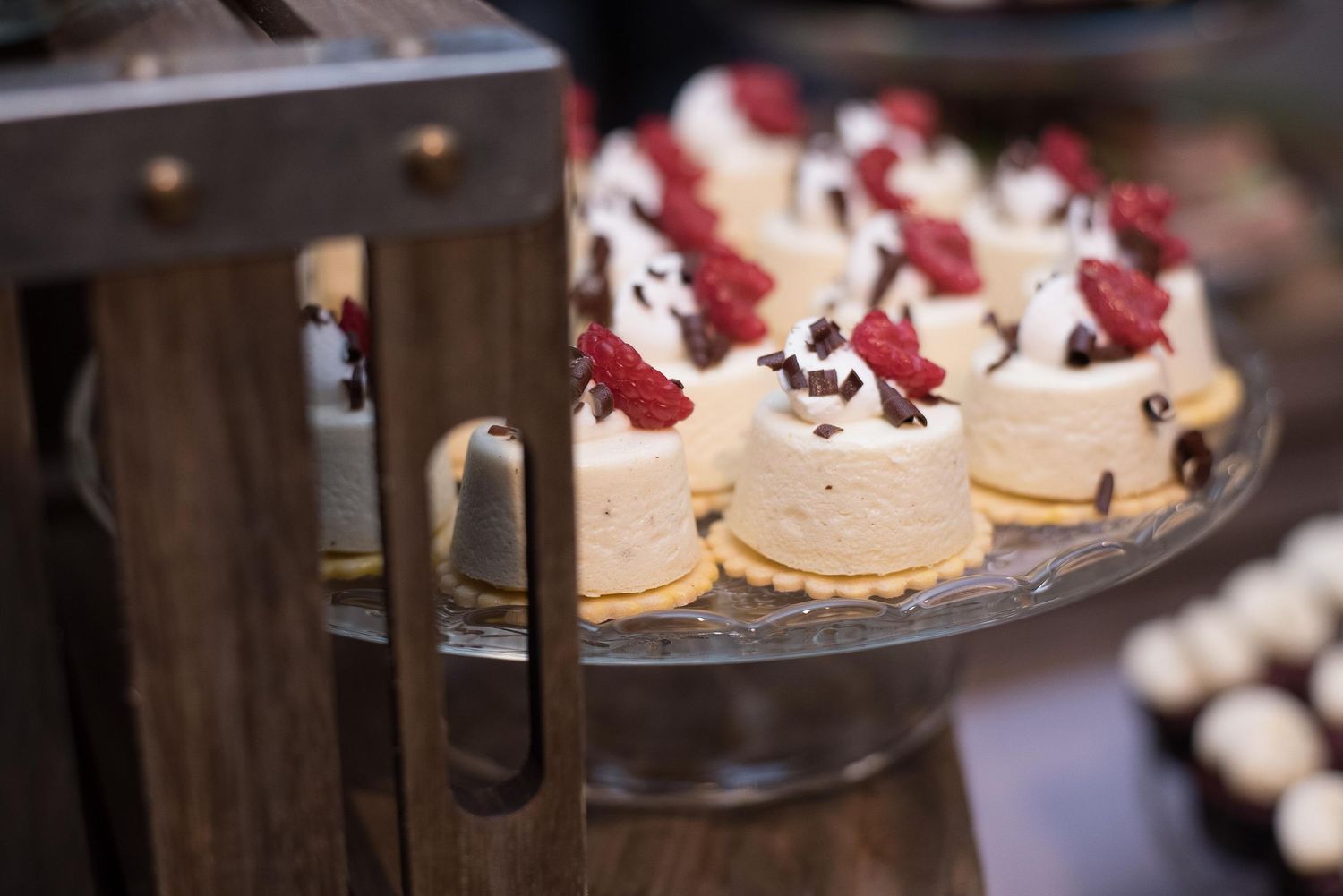 Elegant mini cheesecakes topped with fresh raspberries and chocolate on a glass tiered dessert stand.