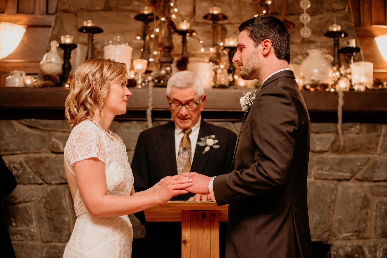 Bride and groom exchange vows at rustic wedding ceremony with officiant and candlelit stone fireplace backdrop.