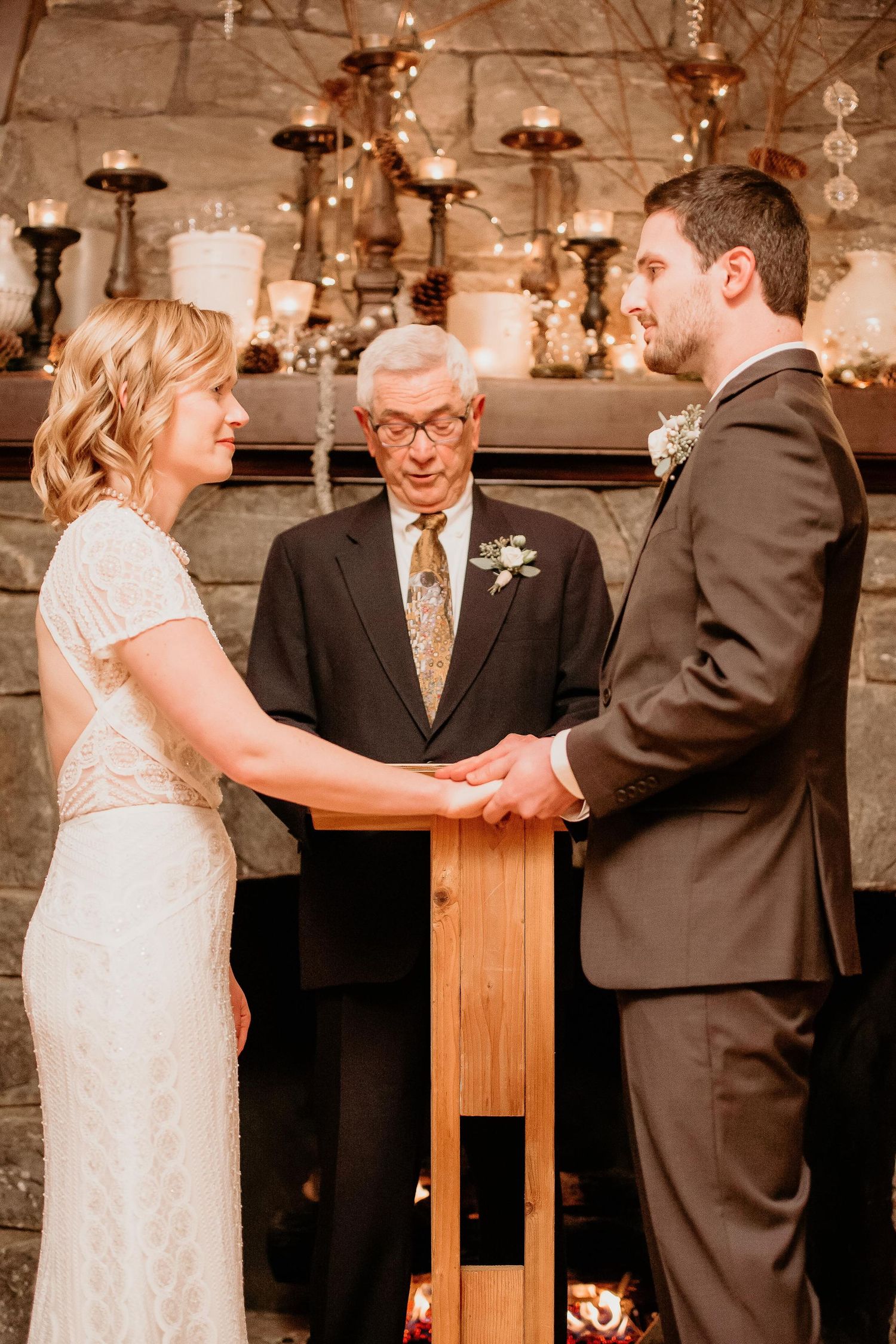 Bride and groom hold hands at rustic fireplace wedding ceremony with officiant and candle decorations.