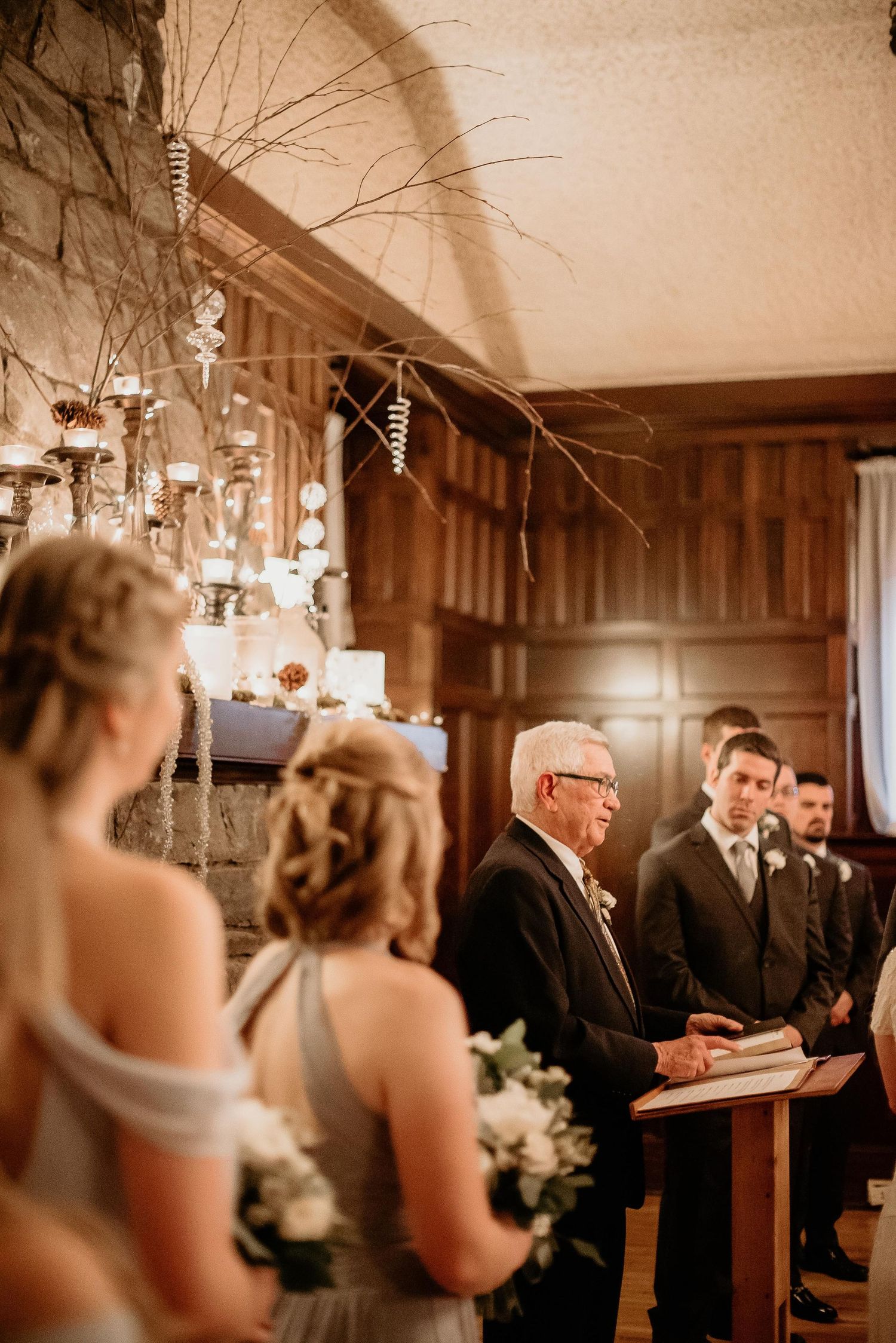 Officiant reads vows at elegant indoor wedding ceremony with wood paneling, bridesmaids holding white bouquets.