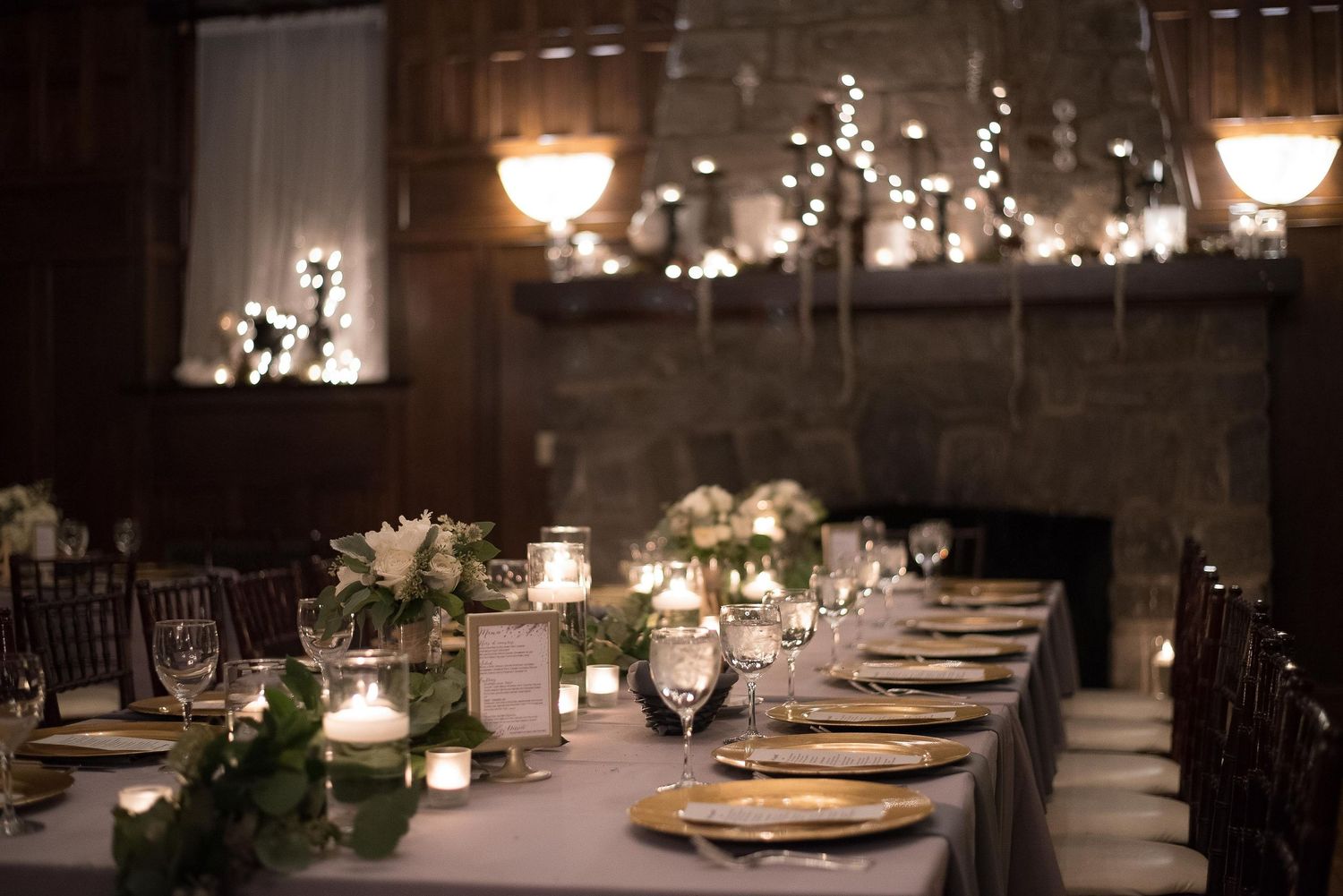 Elegant wedding reception table with gold chargers, candles, and greenery centerpieces in a warmly lit venue.
