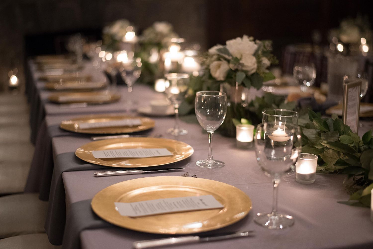 Elegant wedding reception table with gold charger plates, white floral centerpieces, and glowing candles on gray linen.