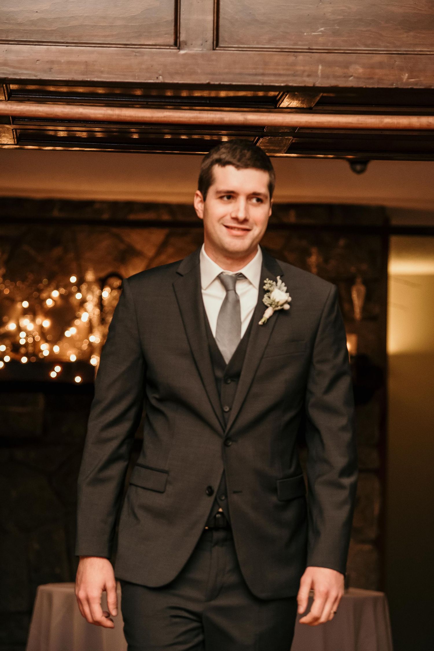 Groom in dark charcoal three-piece suit with gray tie and white boutonniere at elegant wedding reception.