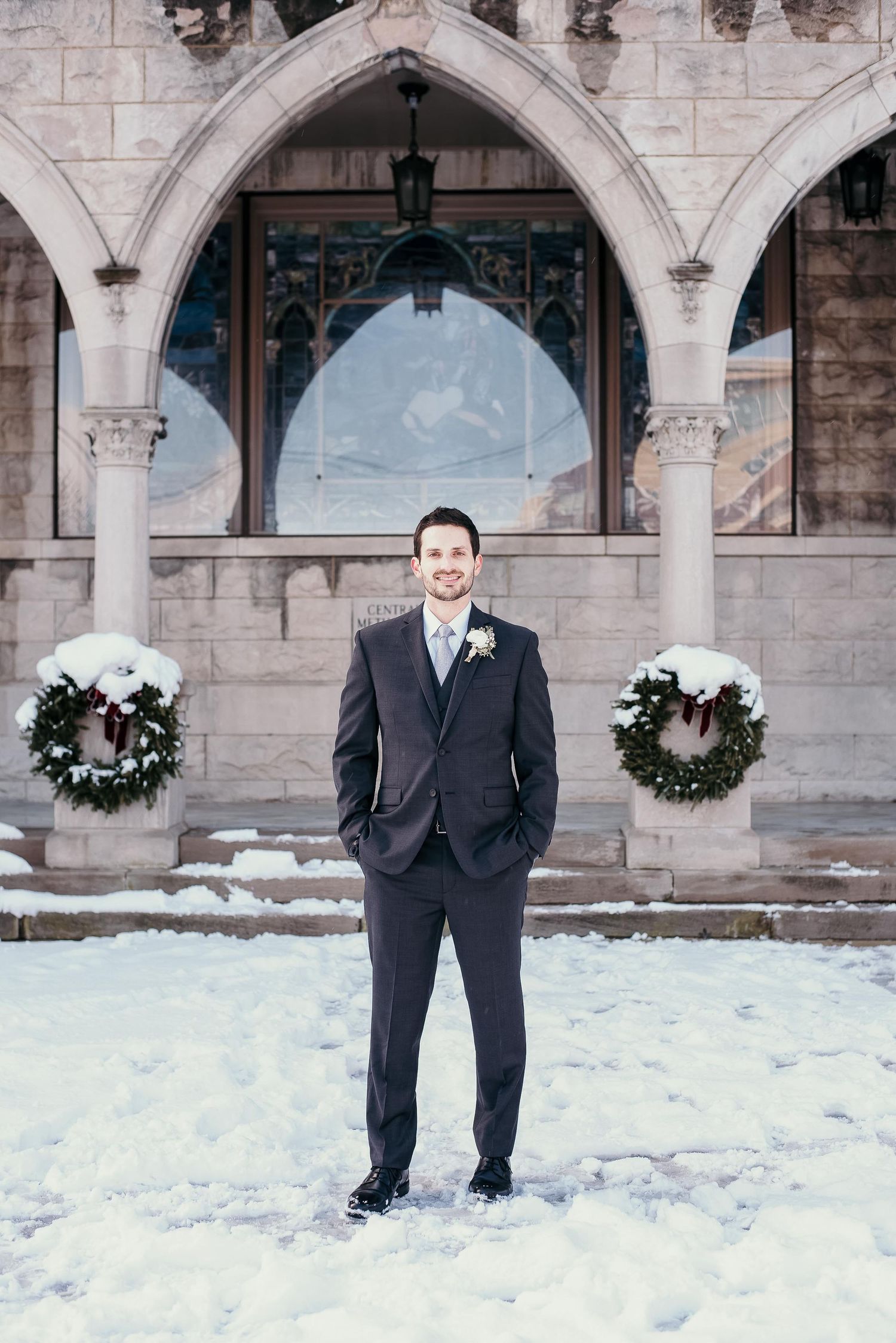 Groom in dark suit stands in snow before Gothic stone building with Christmas wreaths on columns.