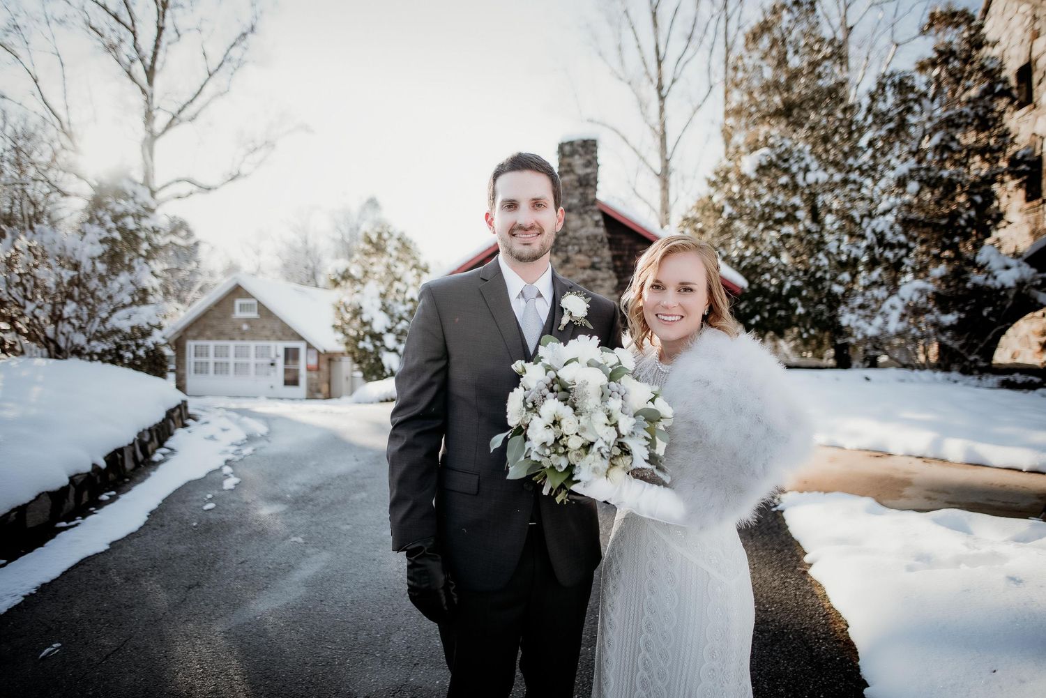 Bride and groom pose together in snow-covered winter wedding setting, bride holding white bouquet wearing fur wrap.
