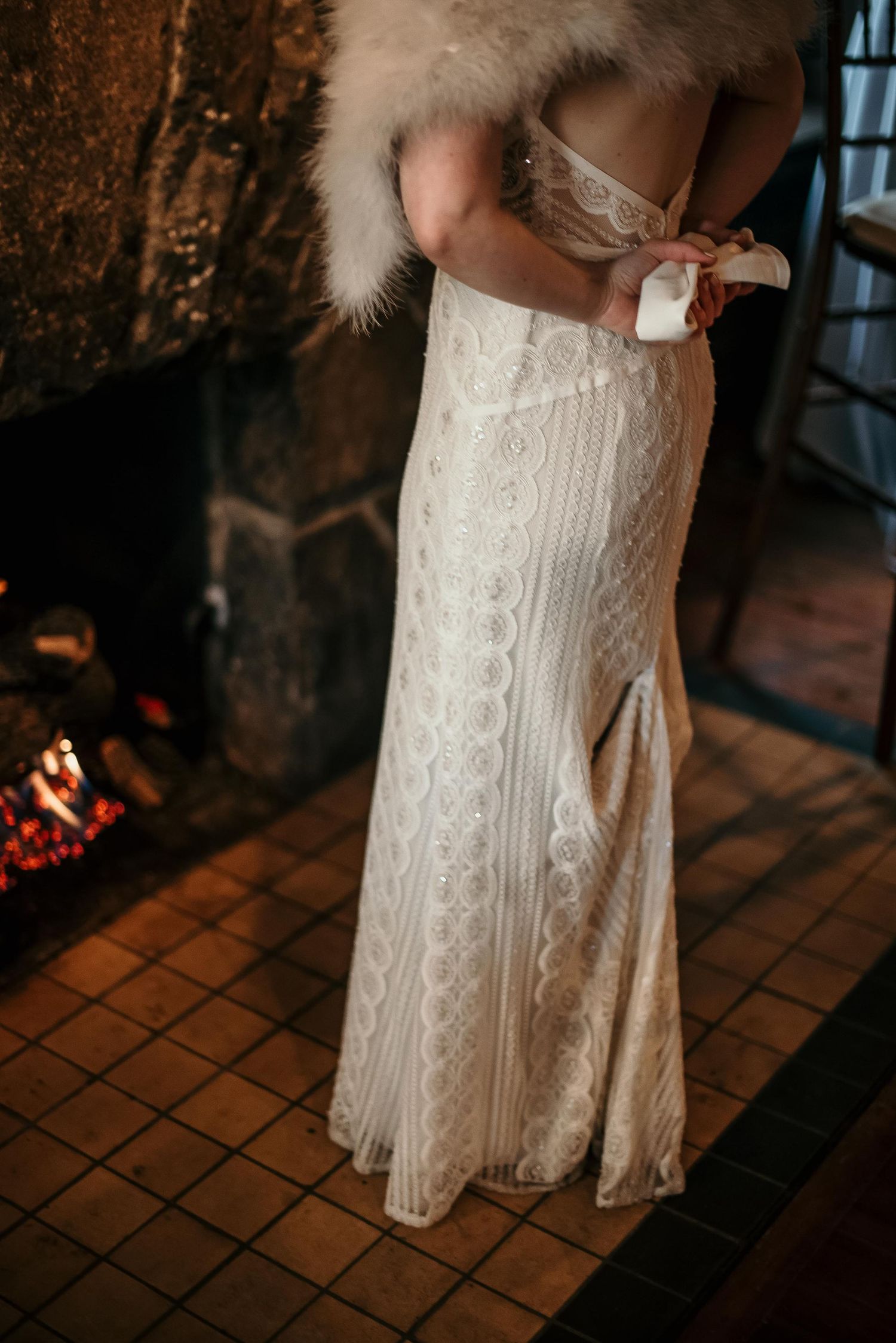 Bride in elegant lace wedding gown with fur stole stands before a glowing fireplace.