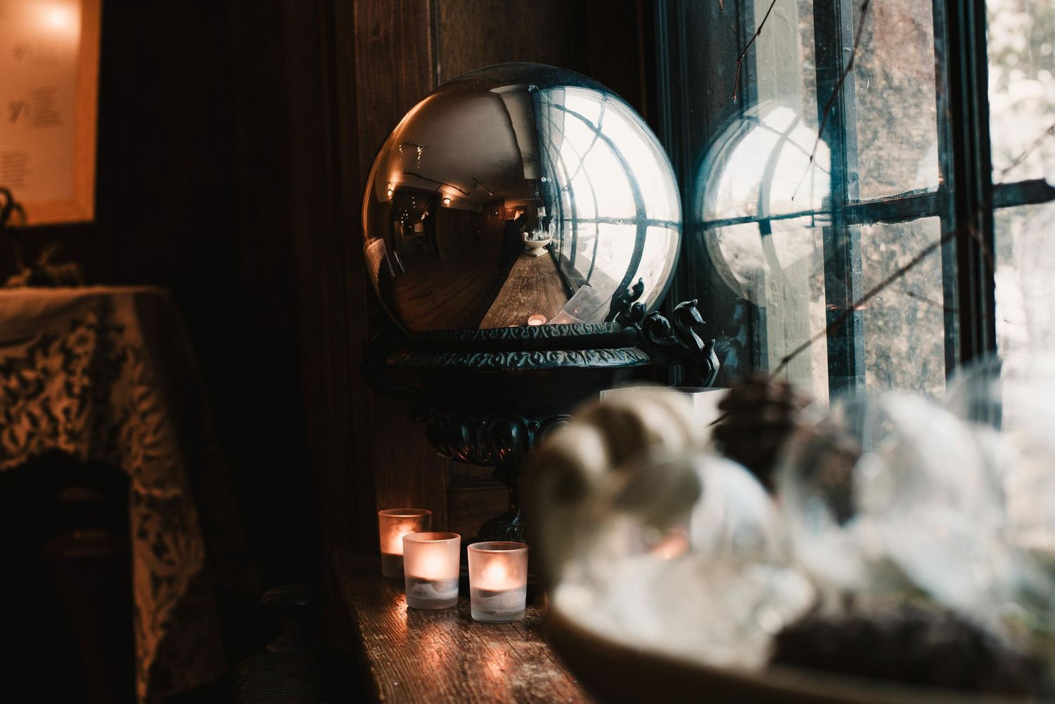 Moody dark interior with glowing candles, crystal ball on ornate stand, and white flowers by a vintage window.