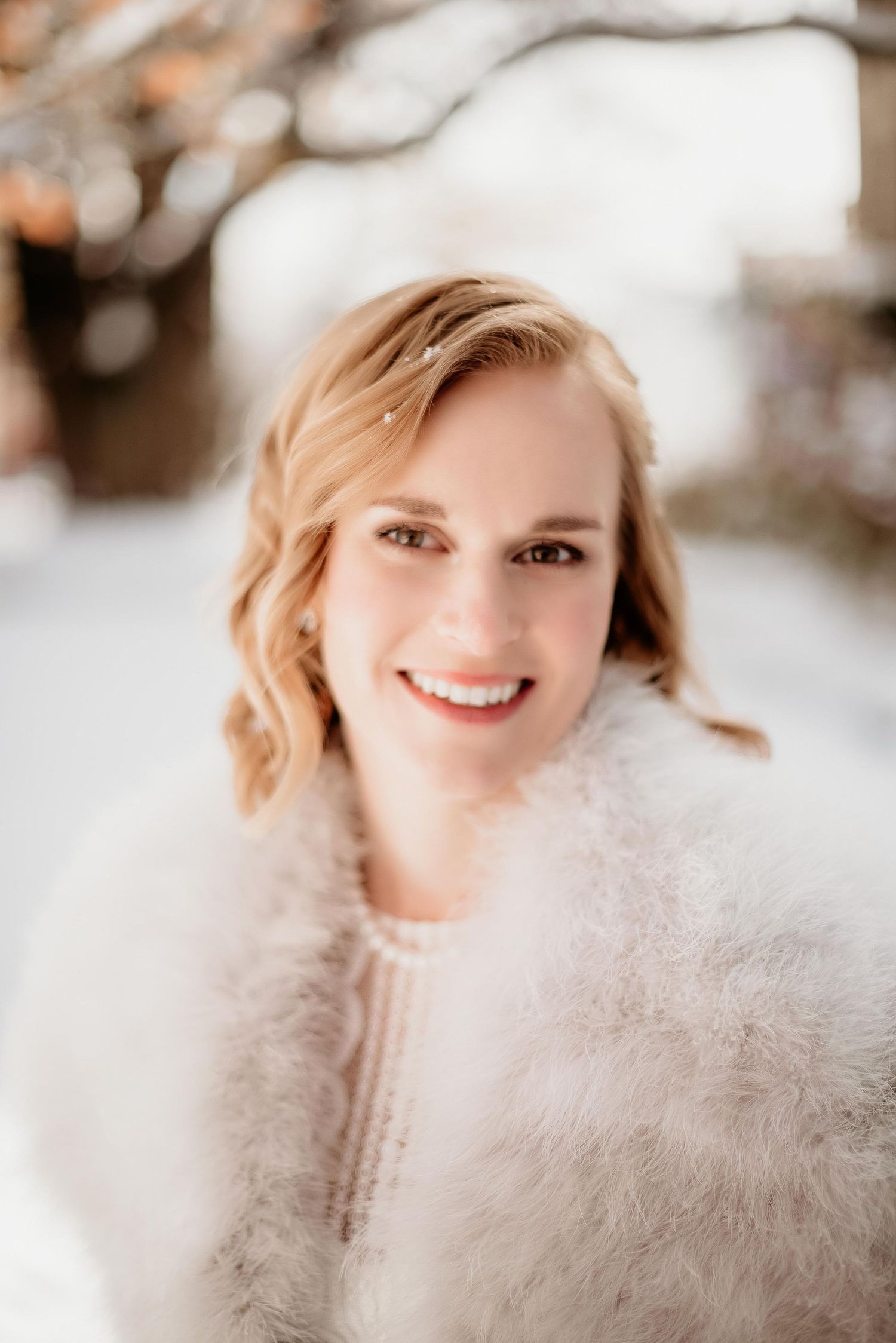 Smiling bride in white feather wrap and pearl necklace with hair accessory poses outdoors in snowy winter setting.