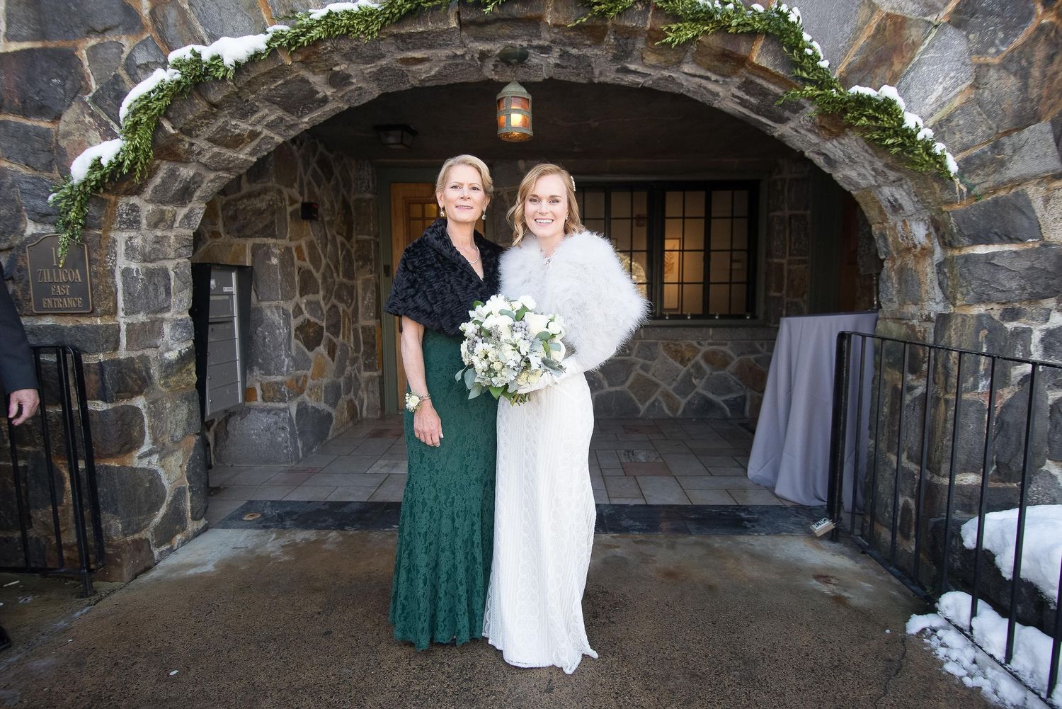 Bride in white gown with fur stole holds bouquet beside woman in green dress under stone arch in winter.