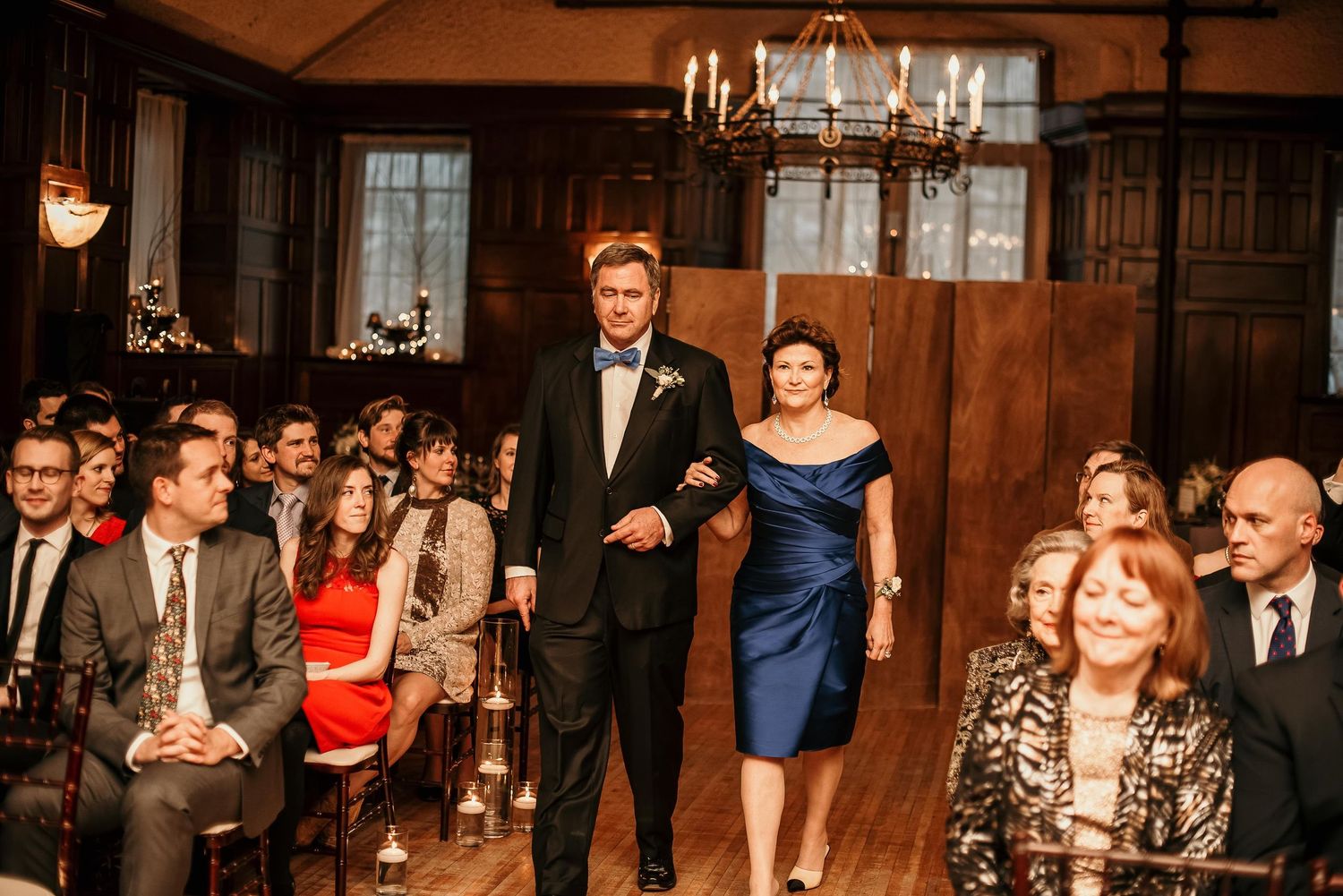 Elegant wedding ceremony with guests seated in a wood-paneled hall as a couple walks down the aisle.