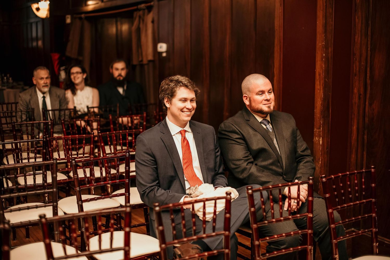 Two men in suits smiling while seated in rows of wooden chairs at an indoor wedding ceremony venue.