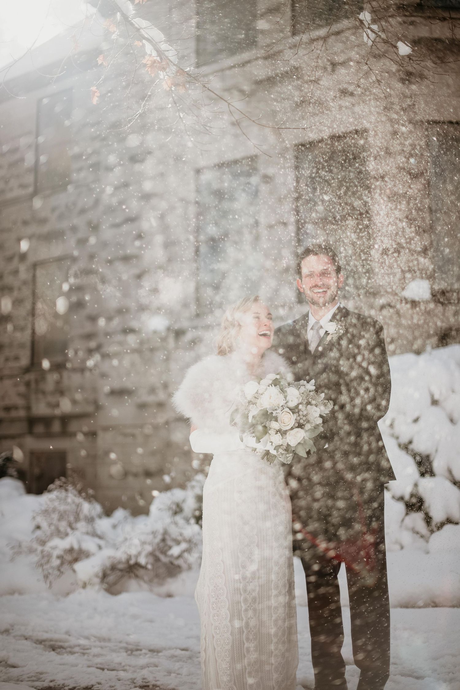 Joyful bride and groom laughing together in a romantic winter snowfall outside a stone building on their wedding day.