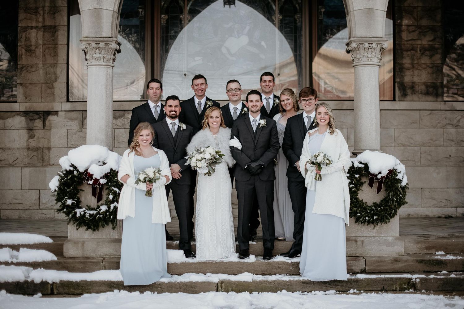 Winter wedding party poses on snowy church steps, bridesmaids in white gowns, groomsmen in gray suits, flanked by wreaths.