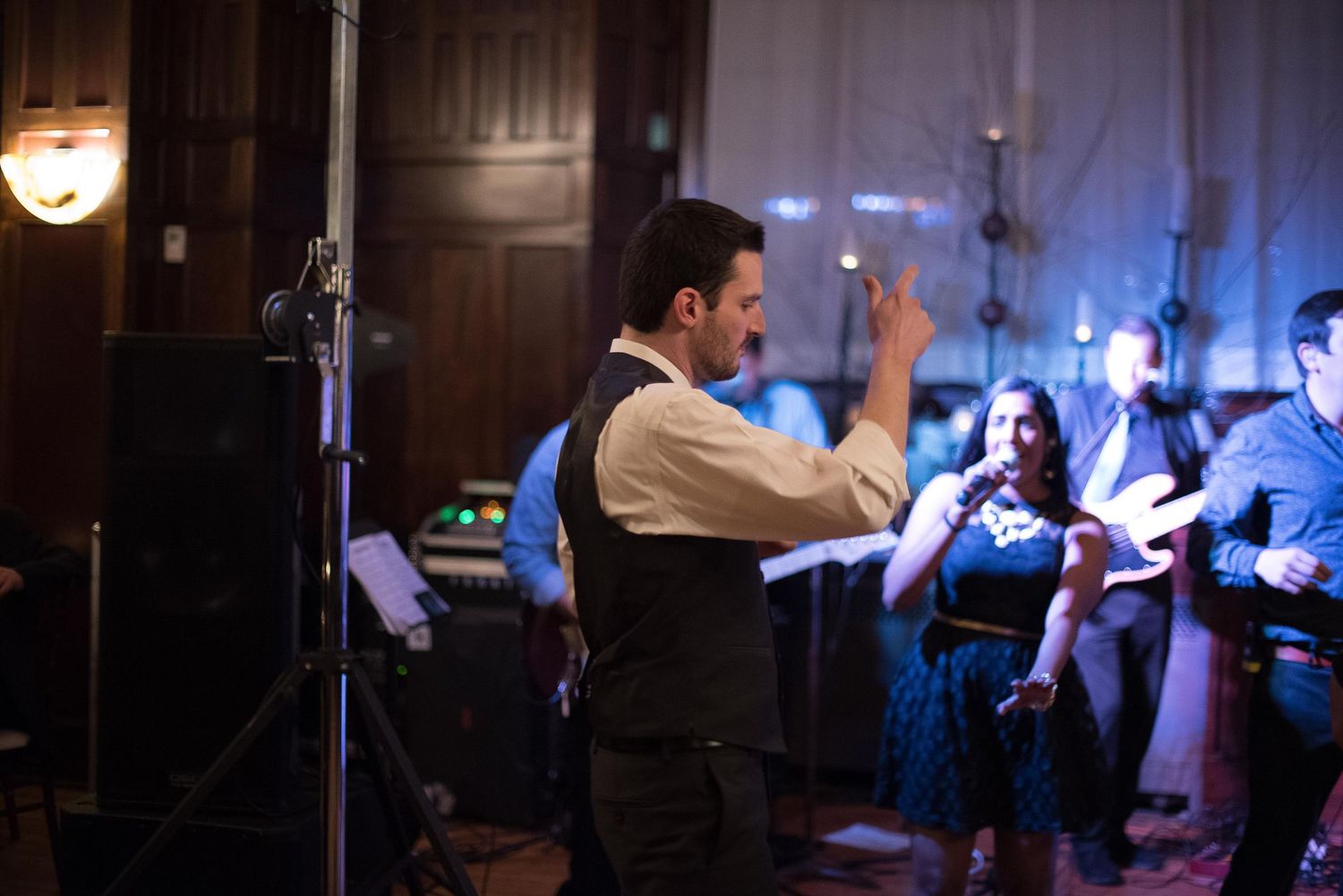 Man in vest conducting a live band performance at an elegant indoor event with musicians in background.
