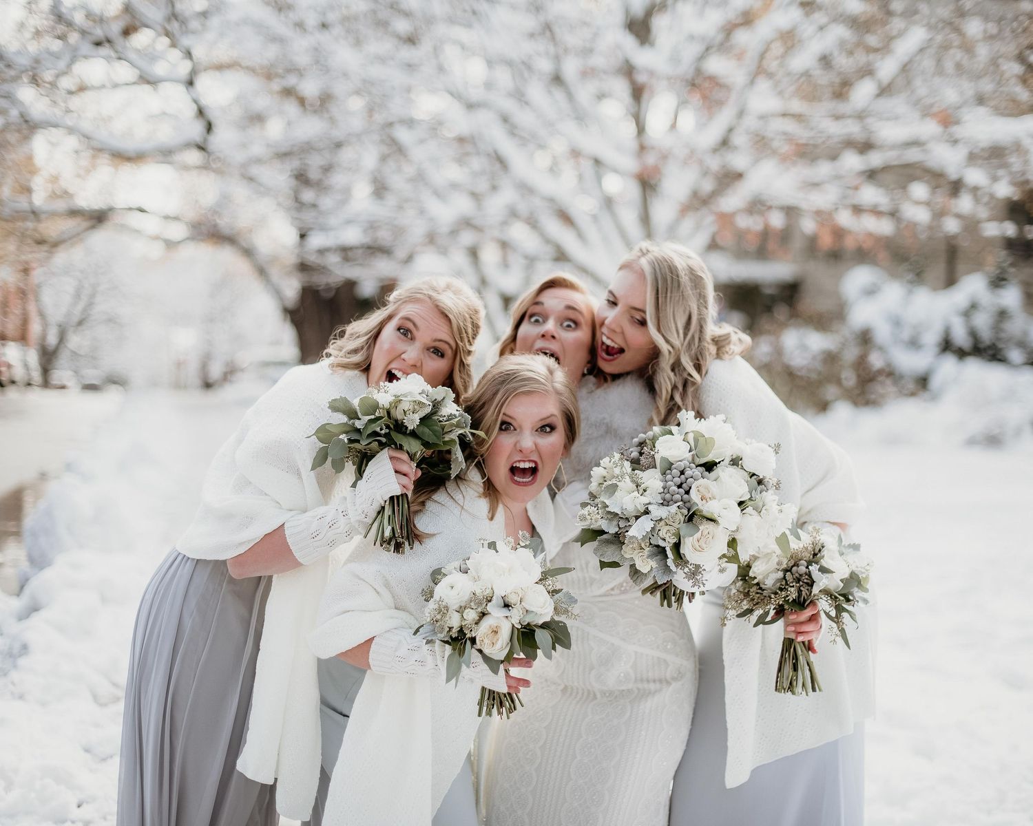 Bride and bridesmaids in white wraps holding bouquets laughing together in a snowy winter wedding outdoor setting.