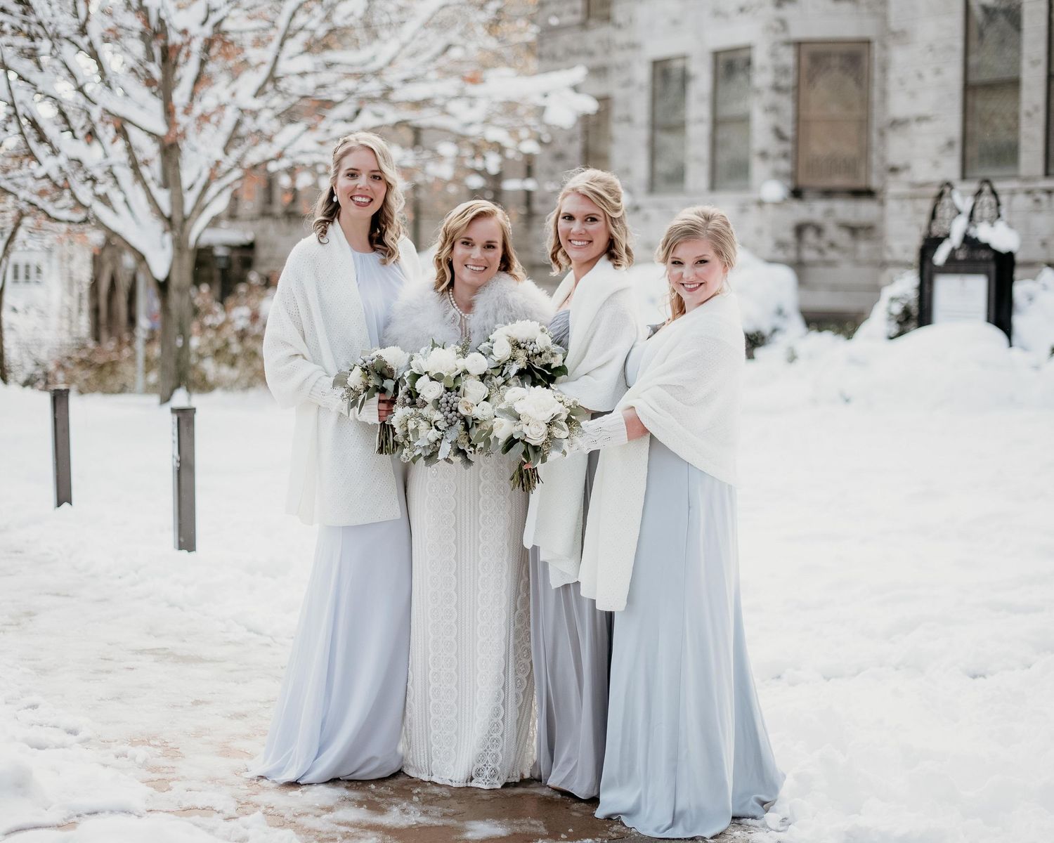 Bride and bridesmaids in light blue gowns with white wraps holding bouquets in a snowy winter wedding setting.