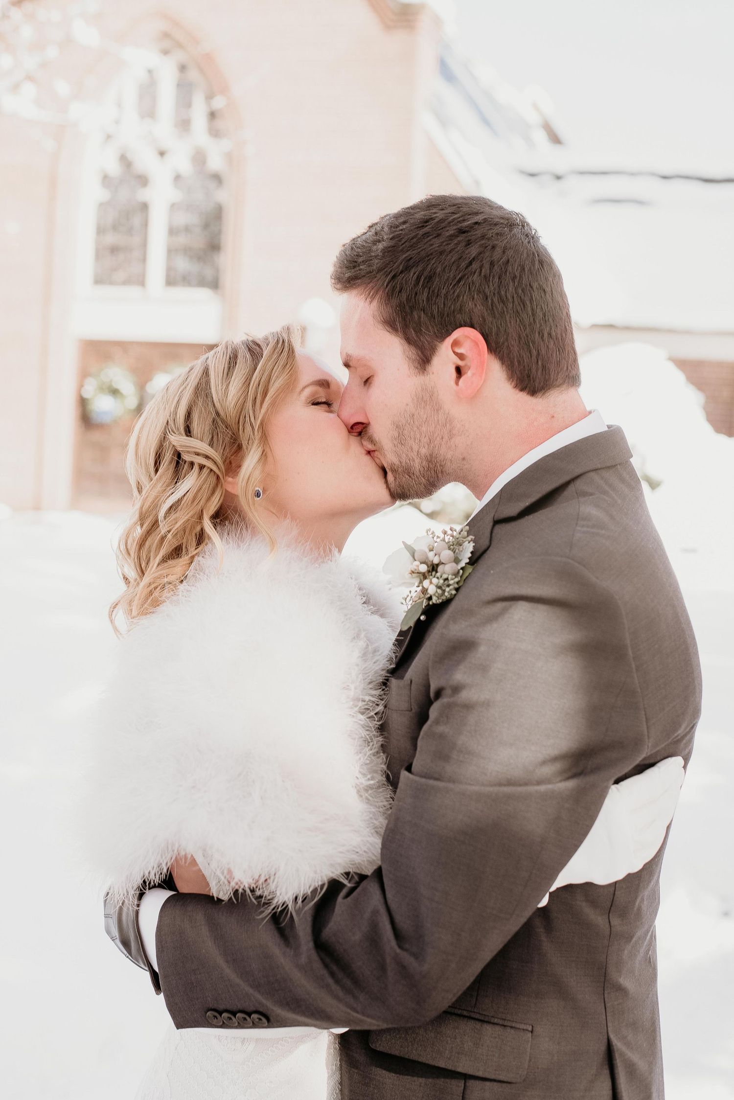 Bride and groom share a romantic winter kiss outdoors, bride wearing a white fur wrap, groom in a gray suit.
