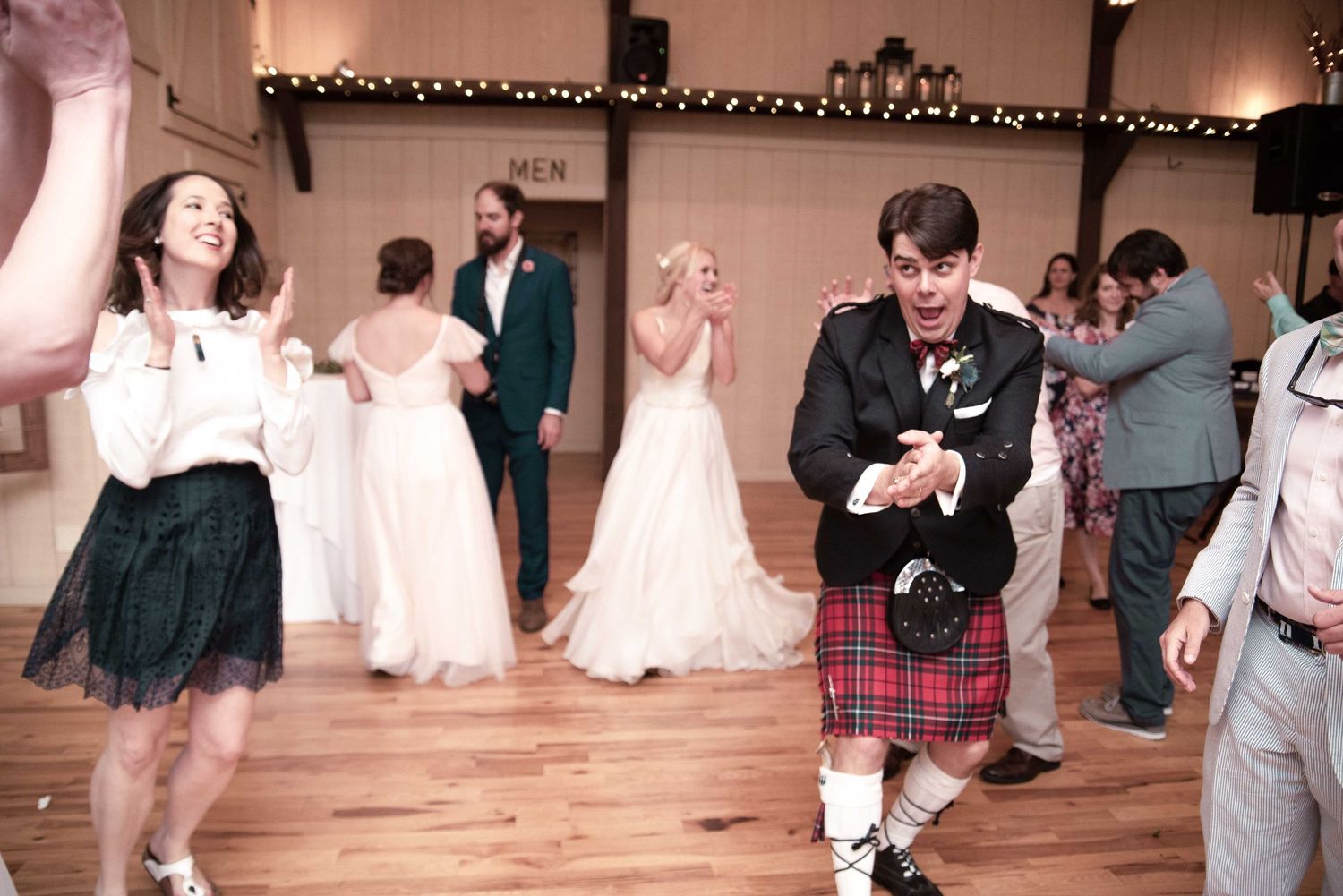 Wedding guests dancing joyfully, man in red tartan kilt celebrating on wooden dance floor at rustic barn reception.