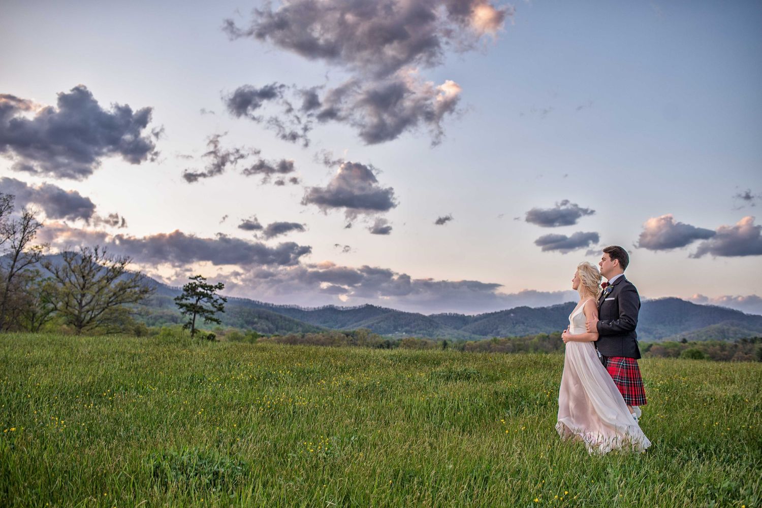 Couple embraces in lush green field at sunset with mountain backdrop and dramatic cloudy sky.