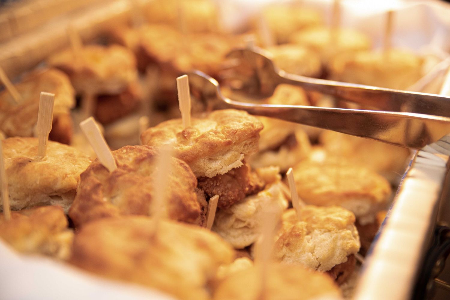 Golden baked appetizers with toothpicks served in a tray, held by silver tongs at a catering event.