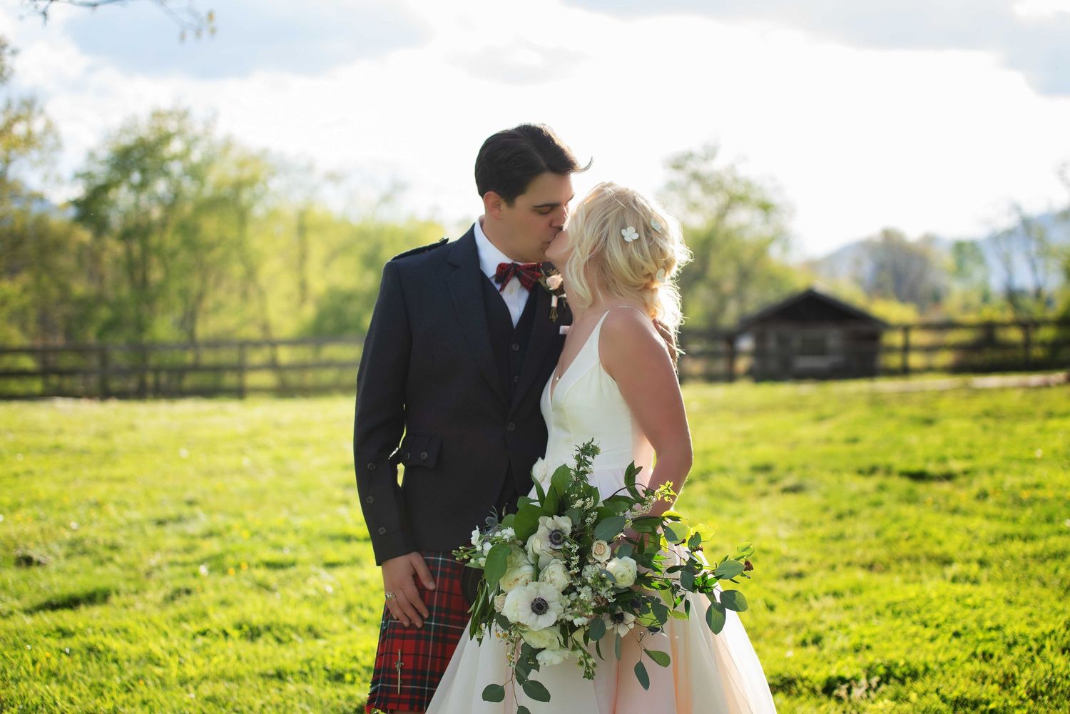 Bride and groom share a romantic moment in a sunlit green field, she holds a lush white floral bouquet, he wears a kilt.