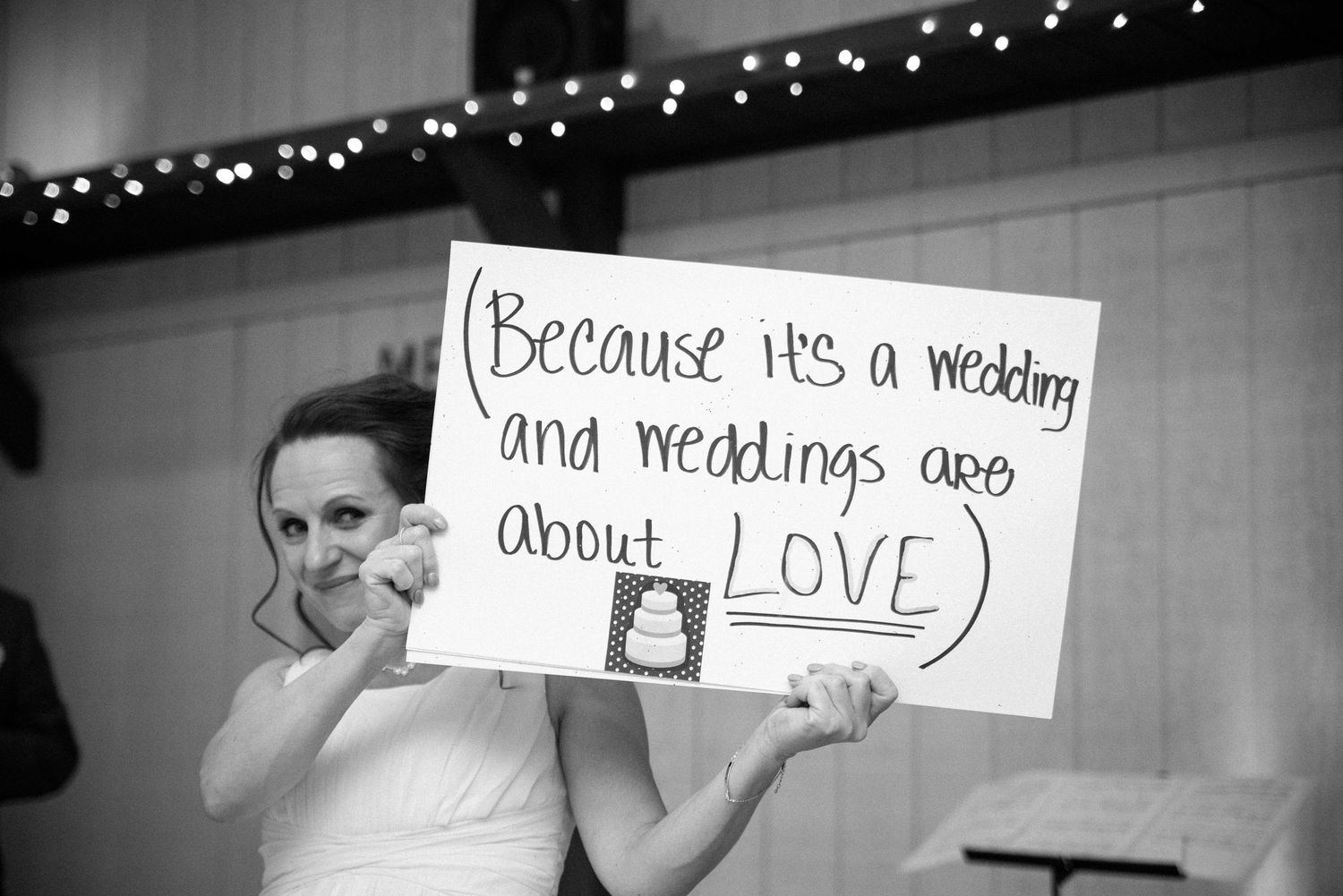 Bride holding sign reading 'Because it's a wedding and weddings are about LOVE' at wedding reception.